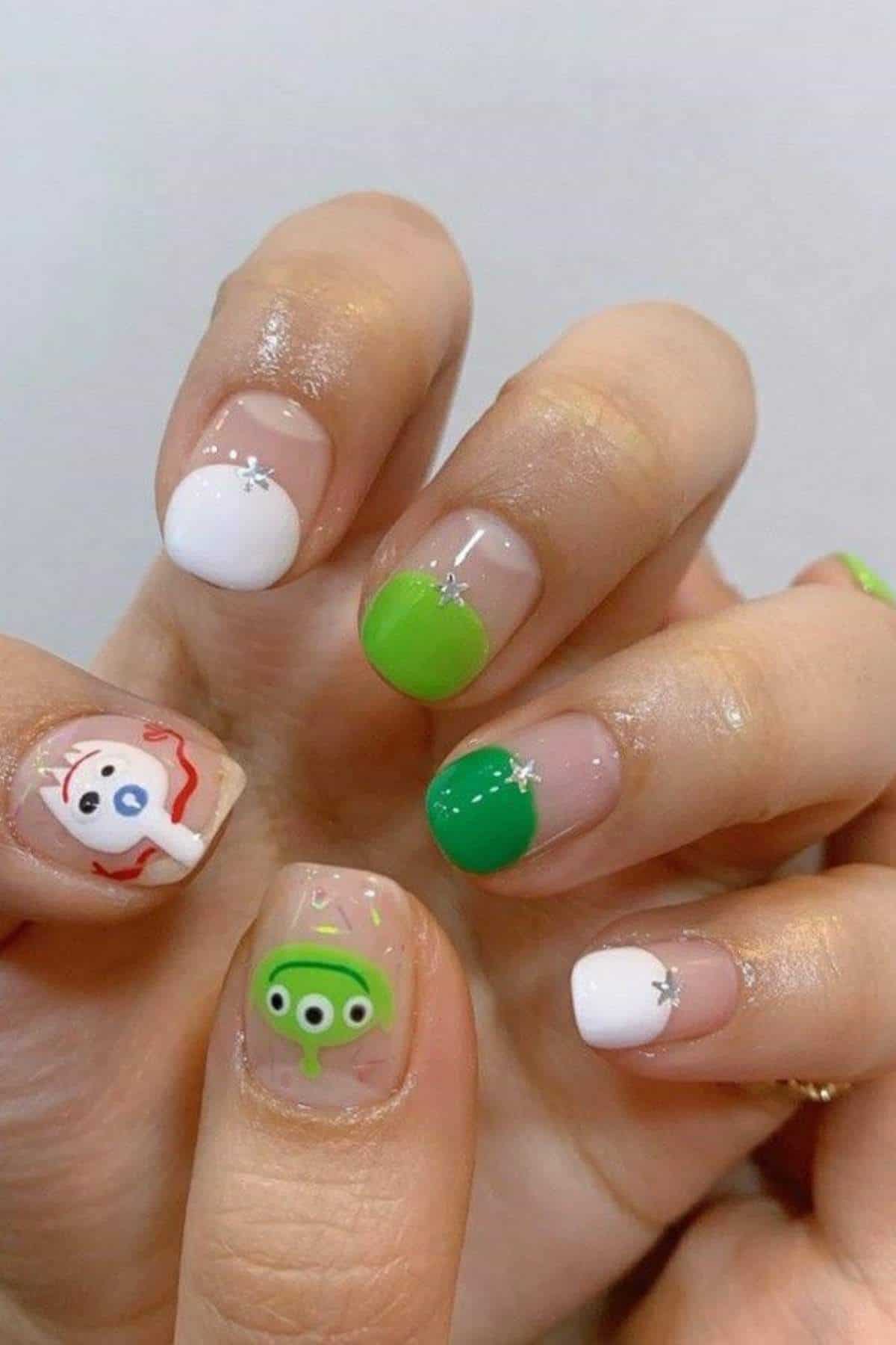 A hand with manicured nails featuring green and white designs, star accents, and cartoon character faces on the thumb and middle finger.