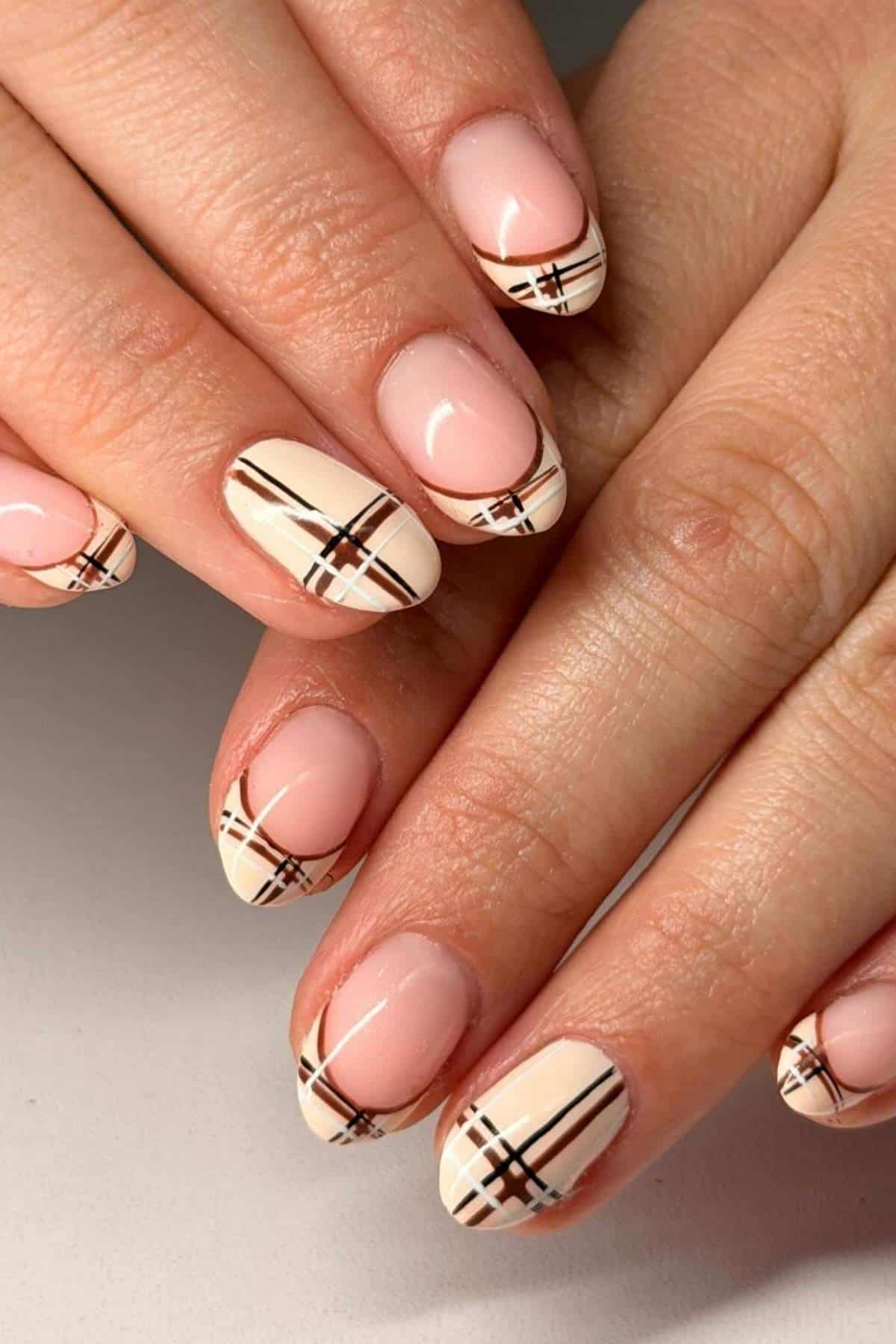 Hands with almond-shaped nails featuring a nude base and white French tips decorated with black and brown plaid lines.