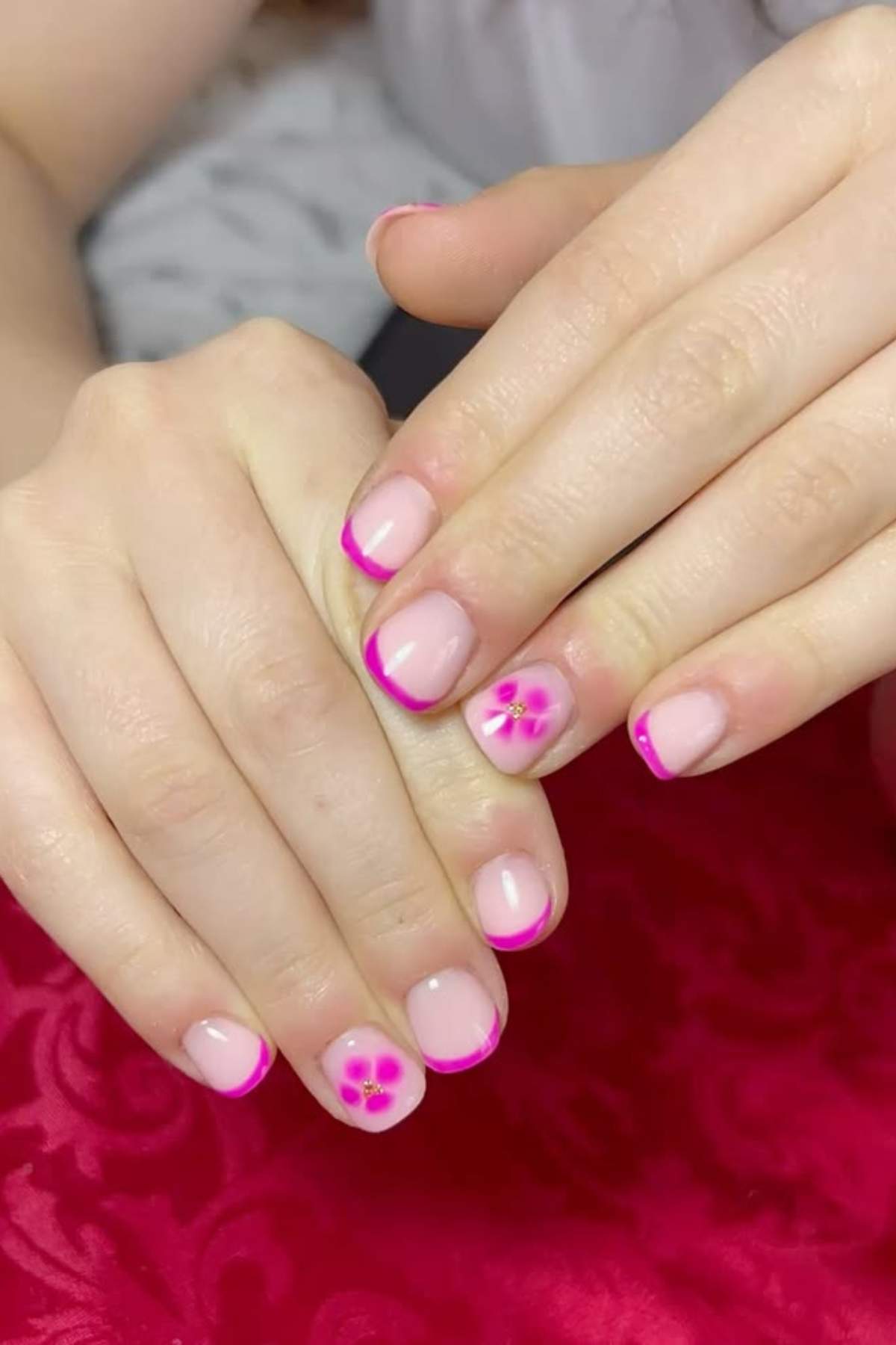 Hands with manicured nails featuring pink French tips and floral designs on the ring fingers, resting on a red surface.