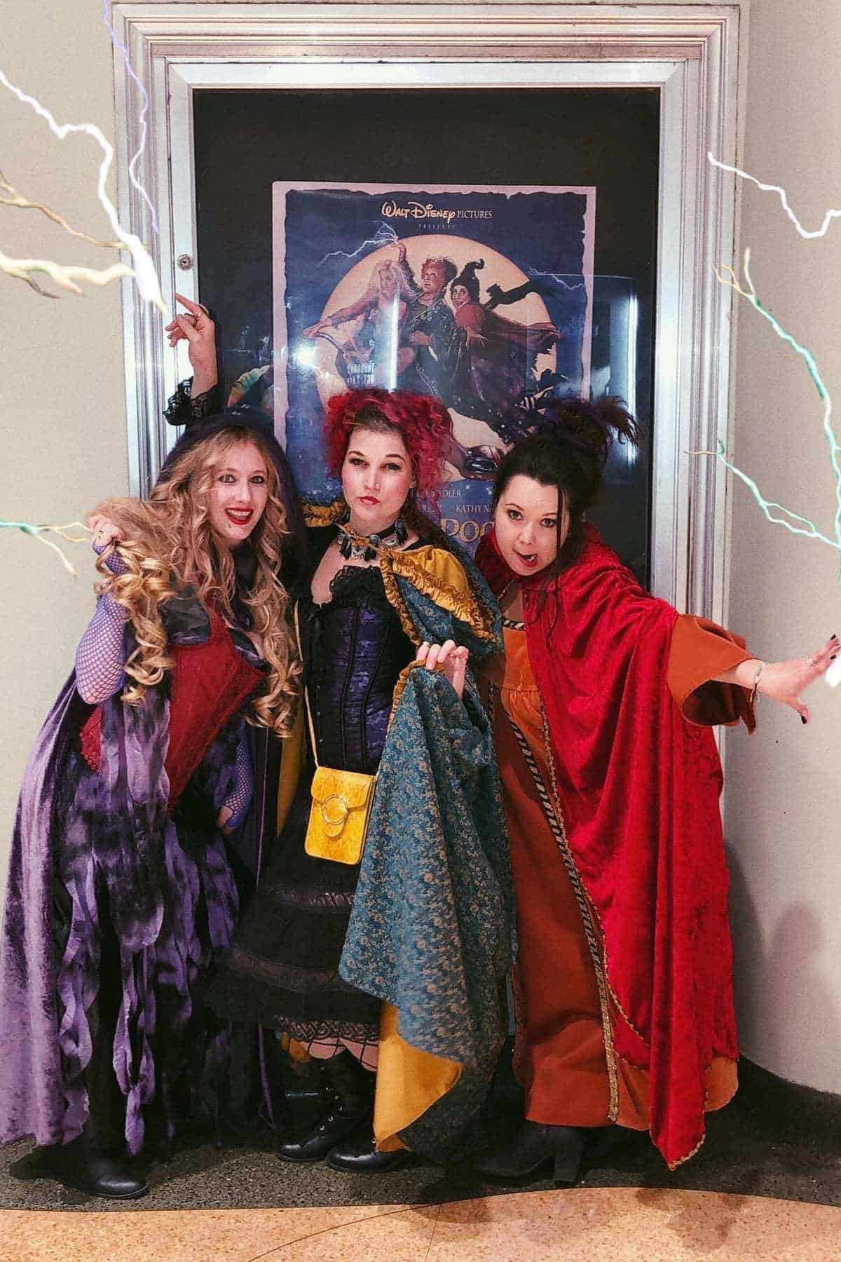 Three women in elaborate Halloween costumes pose in front of a "Hocus Pocus" movie poster, dressed as the Sanderson sisters from the film, with added lightning effects.