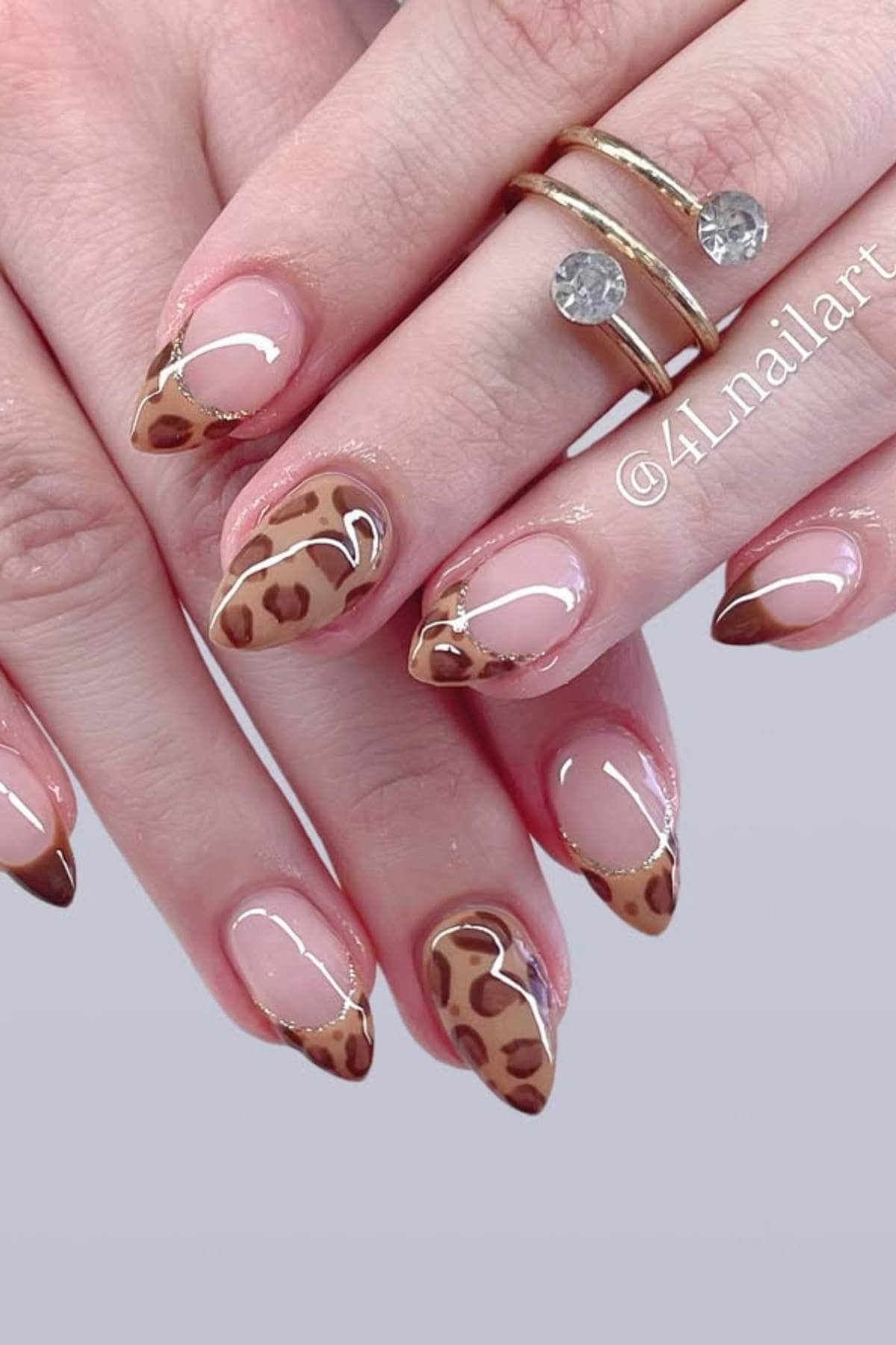 Close-up of hands with almond-shaped nails featuring brown leopard print and French tip nail art, alongside a gold ring with two stones.