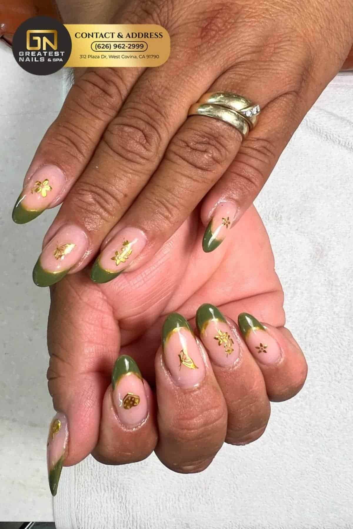 Hands with almond-shaped nails featuring green French tips and gold floral accents, displayed on a white towel. A salon contact sticker appears at the top of the image.