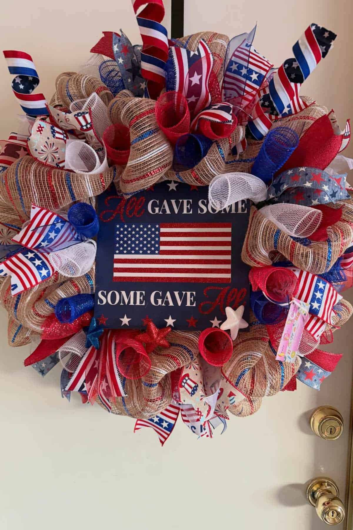 Patriotic wreath with red, white, and blue ribbons, stars, and stripes, featuring a sign that reads "All gave some, some gave all" with an American flag.