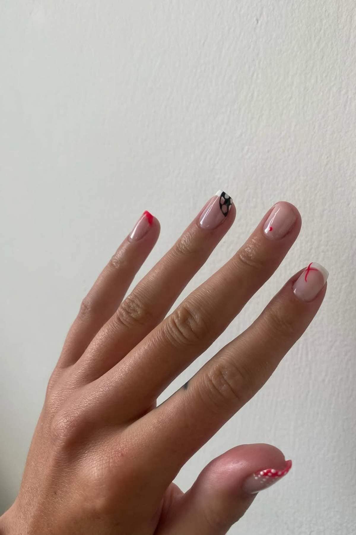 A hand with natural-colored nails featuring minimal red tips and one accent nail with a black abstract design against a plain white background.