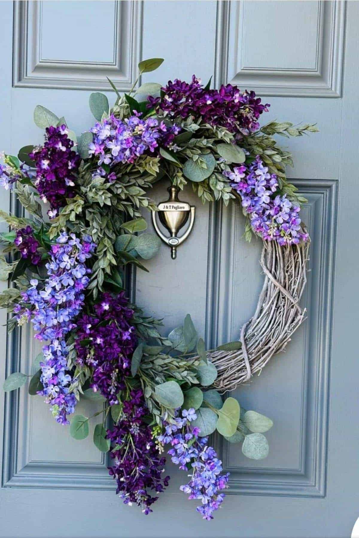 A wreath made of twigs, green leaves, and purple flowers hangs on a gray door, partially covering a silver door knocker.