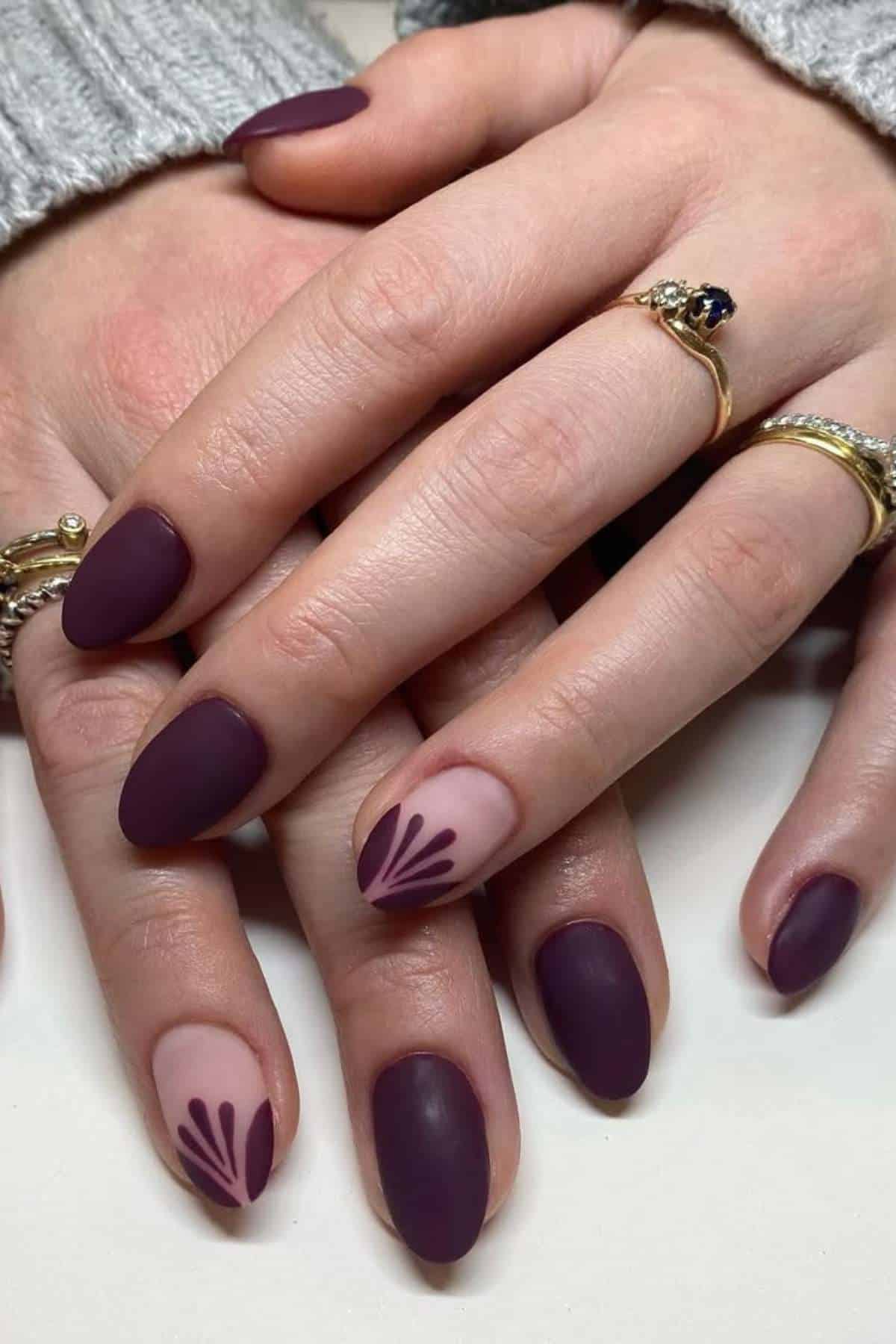 Hands with matte dark purple almond-shaped nails, some featuring nude accents with dark purple floral designs. The person is wearing gold rings and a gray sweater.