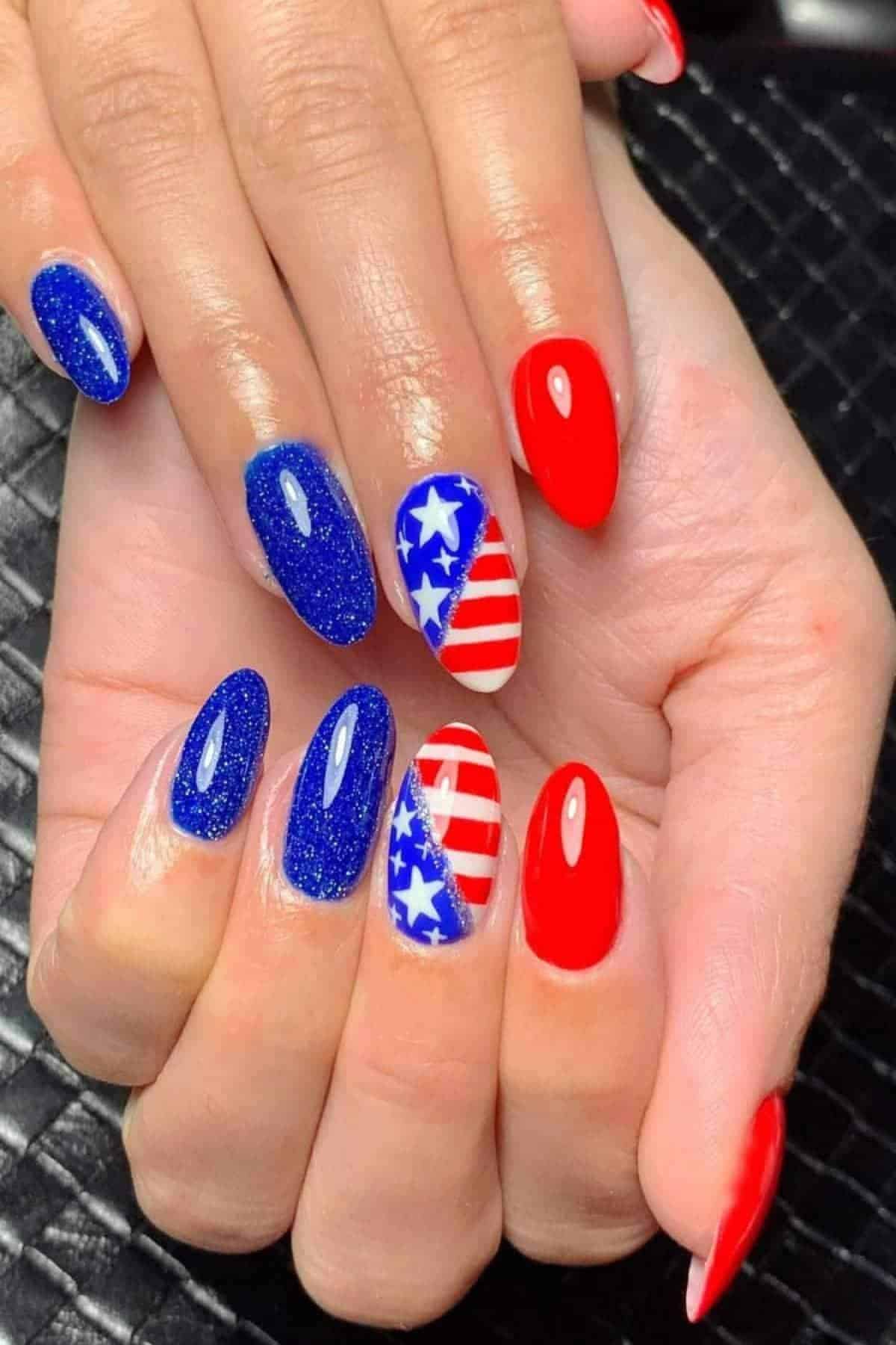 Hands with manicured nails featuring blue glitter, solid red, and accent nails with white stars and red stripes in a patriotic American flag design.