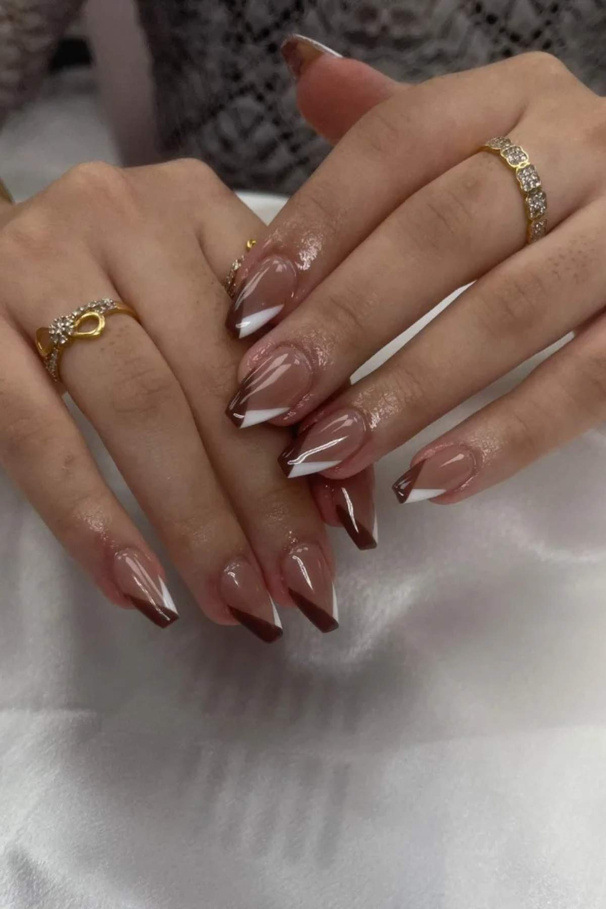 Two hands with long, coffin-shaped nails featuring brown and white French tip designs; the person is wearing gold rings with small stones.