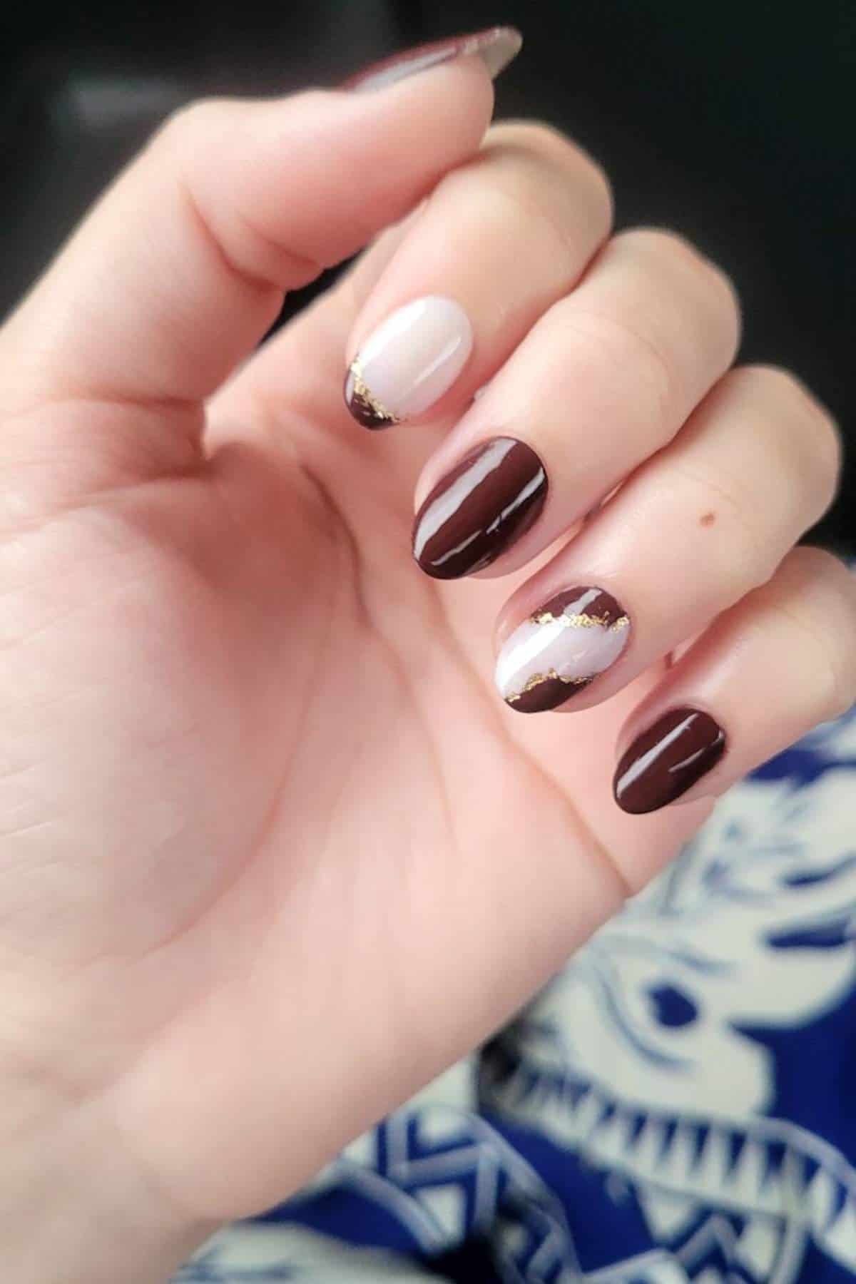 A hand with almond-shaped nails features a brown and nude color scheme, gold accents, and a glossy finish. The background includes blue and white patterned fabric.
