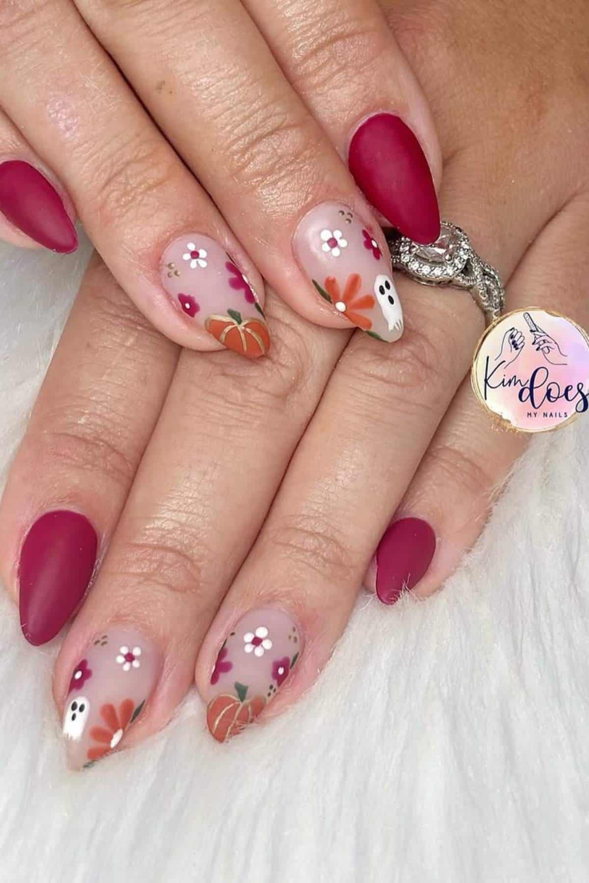 Hands with almond-shaped nails; some are painted matte red while others have fall-themed designs with flowers, pumpkins, and a small ghost on a neutral background.