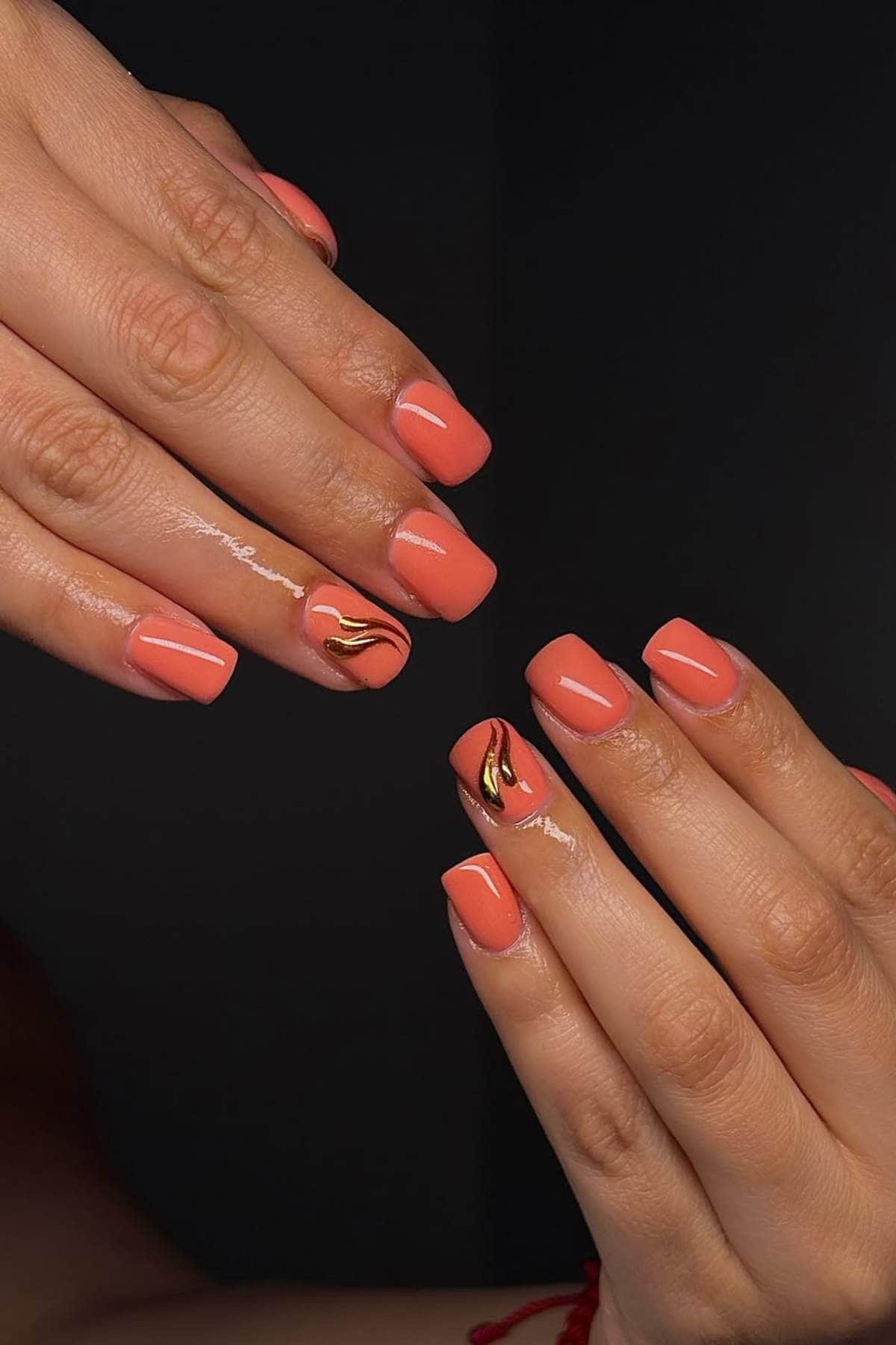 Two hands with short, square, coral-colored nails; some feature gold and black abstract lines as accent nail art.
