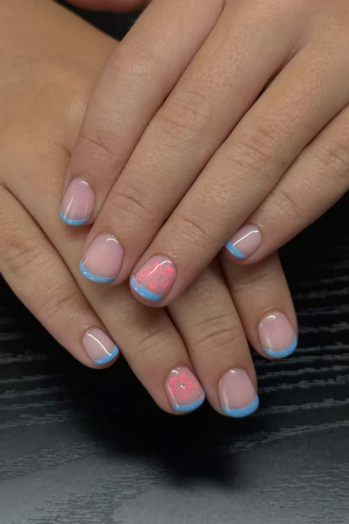 Hands with short, natural nails featuring a light blue French tip; the ring fingernail has a pink heart-shaped design with abstract lines.