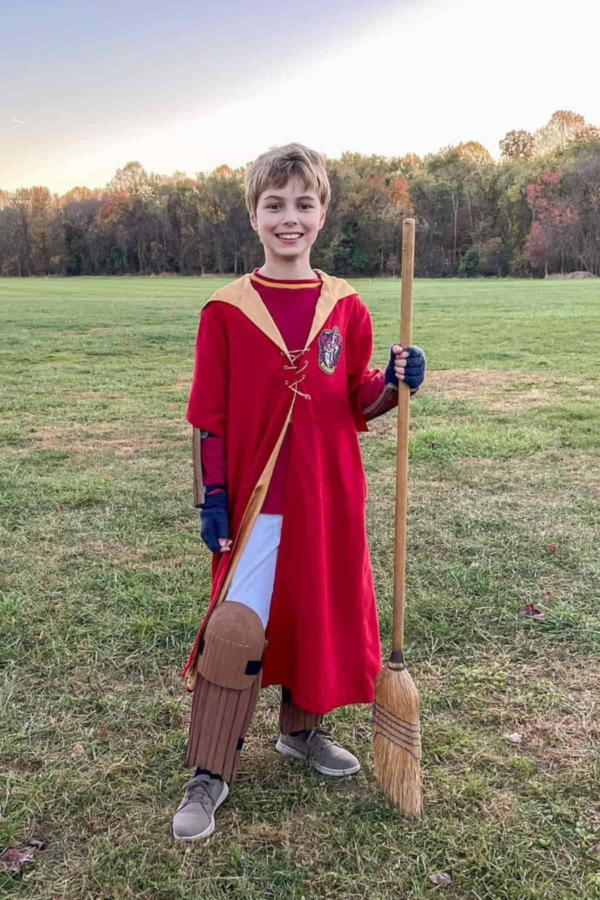 A child dressed in a red and gold robe, holding a broomstick and standing on grass, with trees and a clear sky in the background.