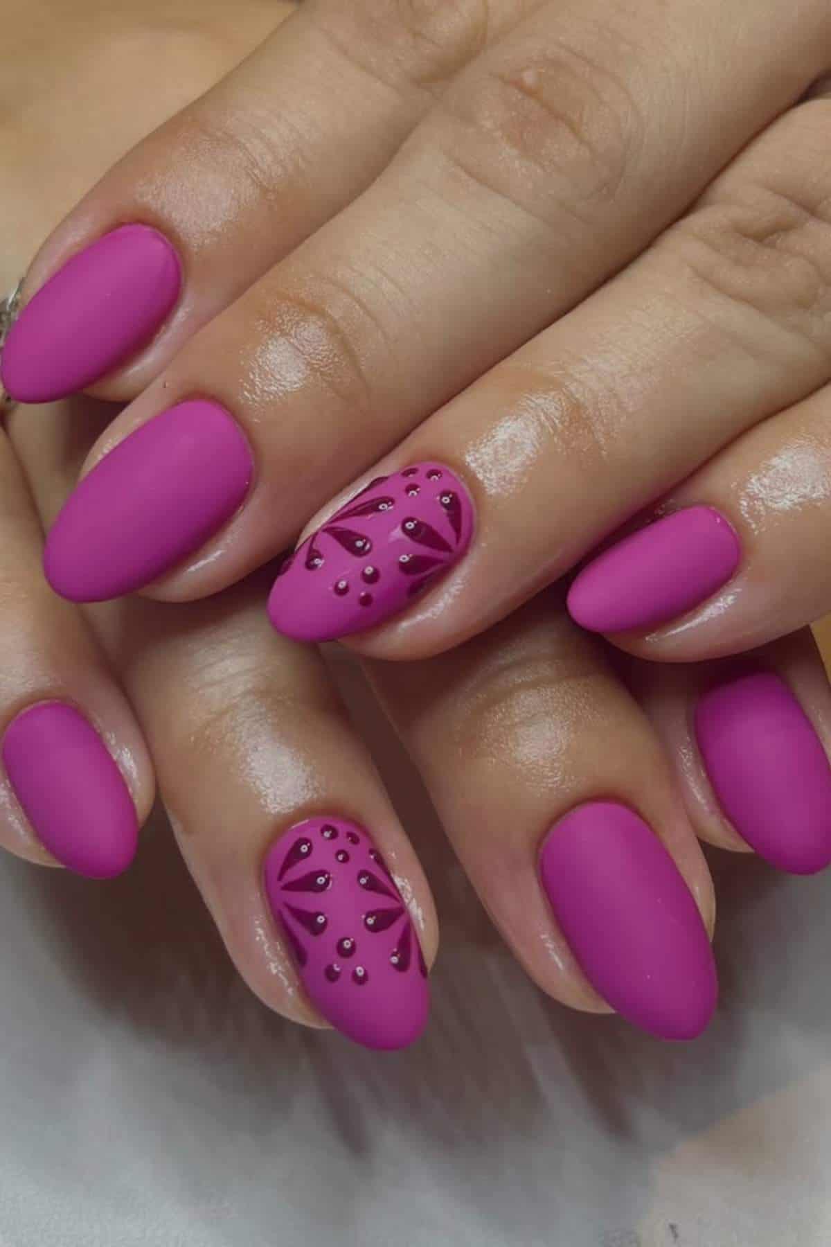 Hands with oval-shaped nails painted matte magenta; ring finger nails feature geometric patterns and small studs as decoration.