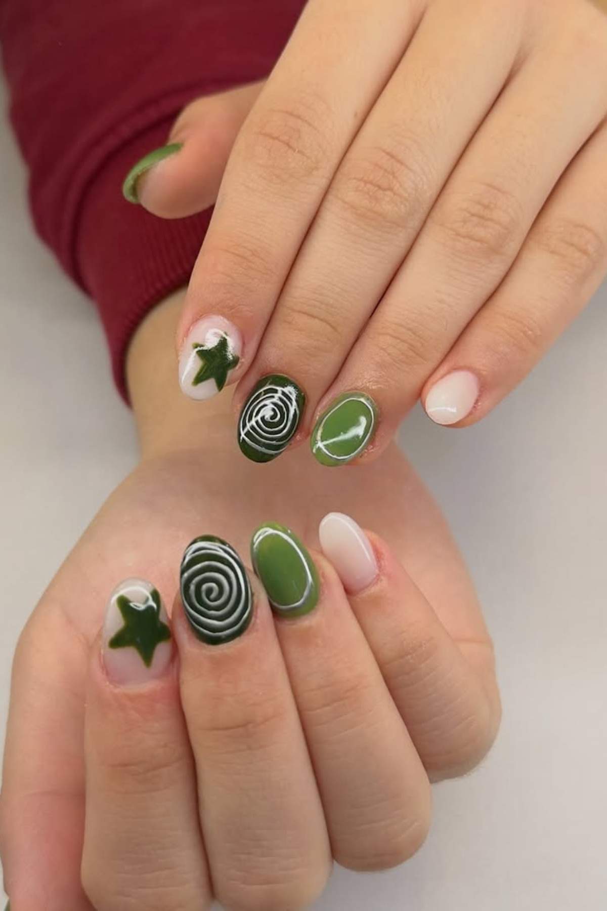 Hands with manicured nails featuring green spiral, star, solid green, and nude designs on short, oval-shaped nails.