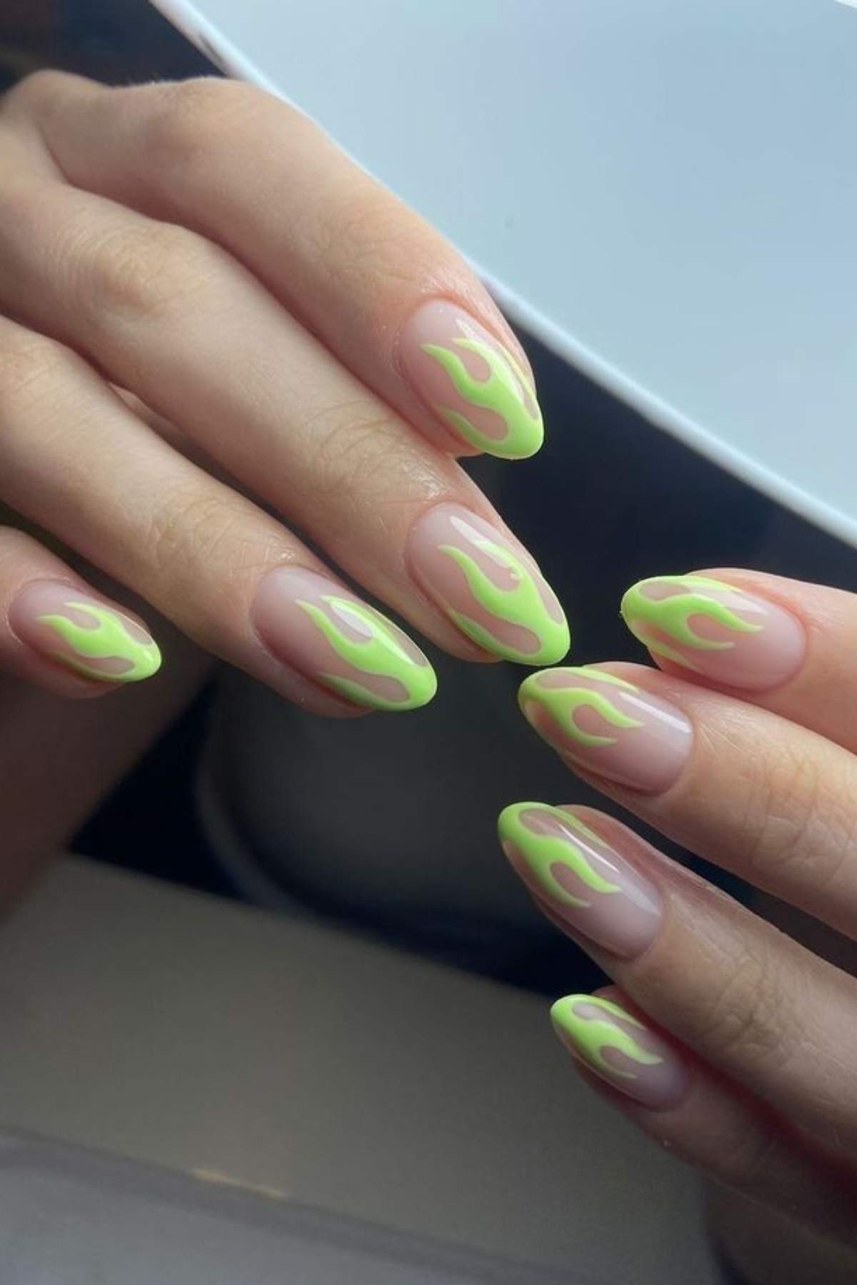 Hands with almond-shaped nails featuring a nude base and neon green flame nail art at the tips.