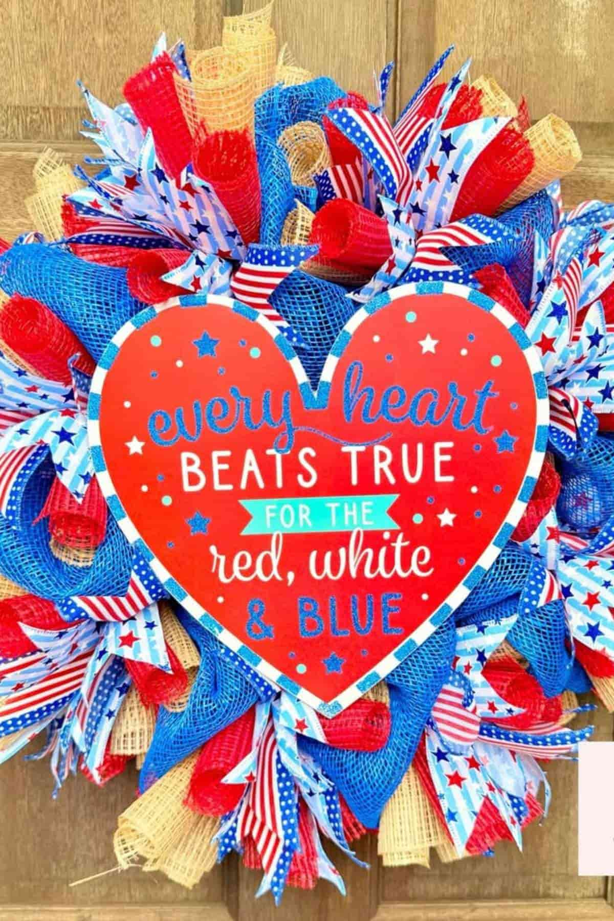 Patriotic wreath decorated with red, white, and blue ribbons, featuring a heart-shaped sign that reads, “every heart BEATS TRUE FOR THE red, white & BLUE.”.