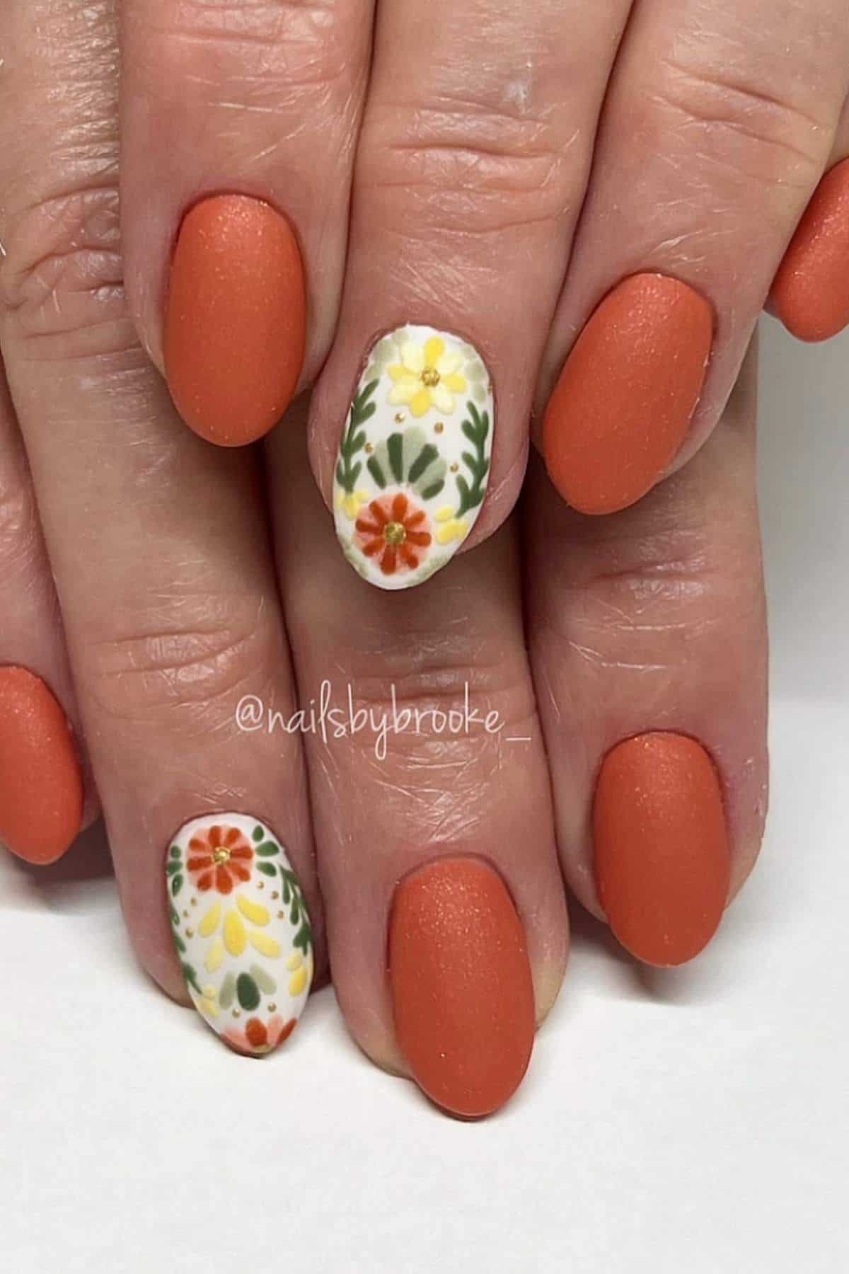 Hands with matte orange nail polish, featuring white accent nails with colorful floral designs on the ring fingers.