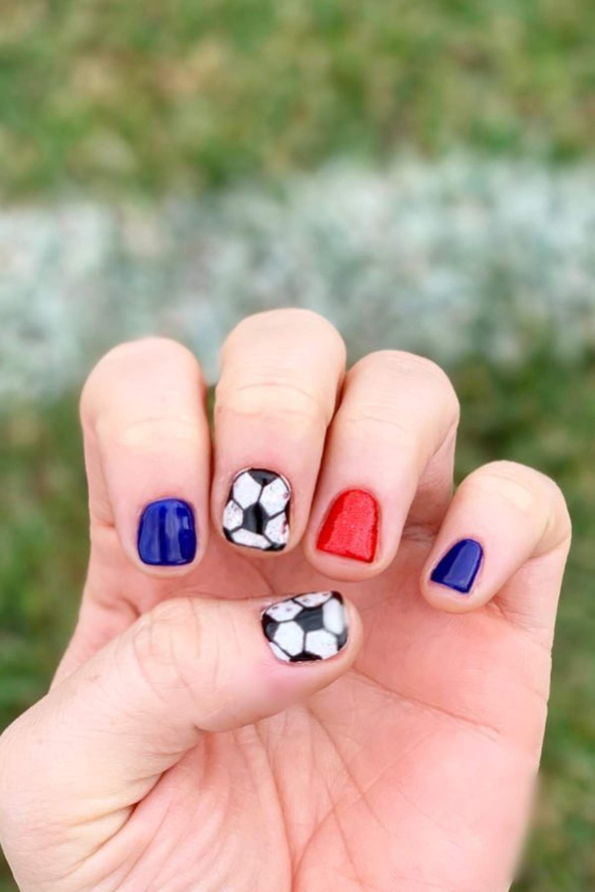 A hand displays nails painted blue, red, and decorated with black and white soccer ball designs, shown outdoors on green grass.