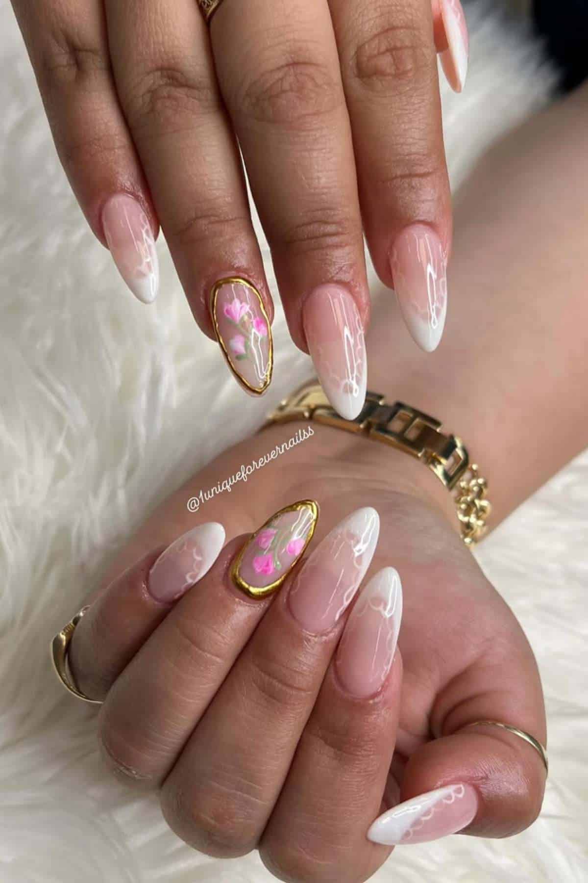 Close-up of hands with almond-shaped nails featuring a pink and white French tip design; one nail on each hand has a floral accent with gold trim.
