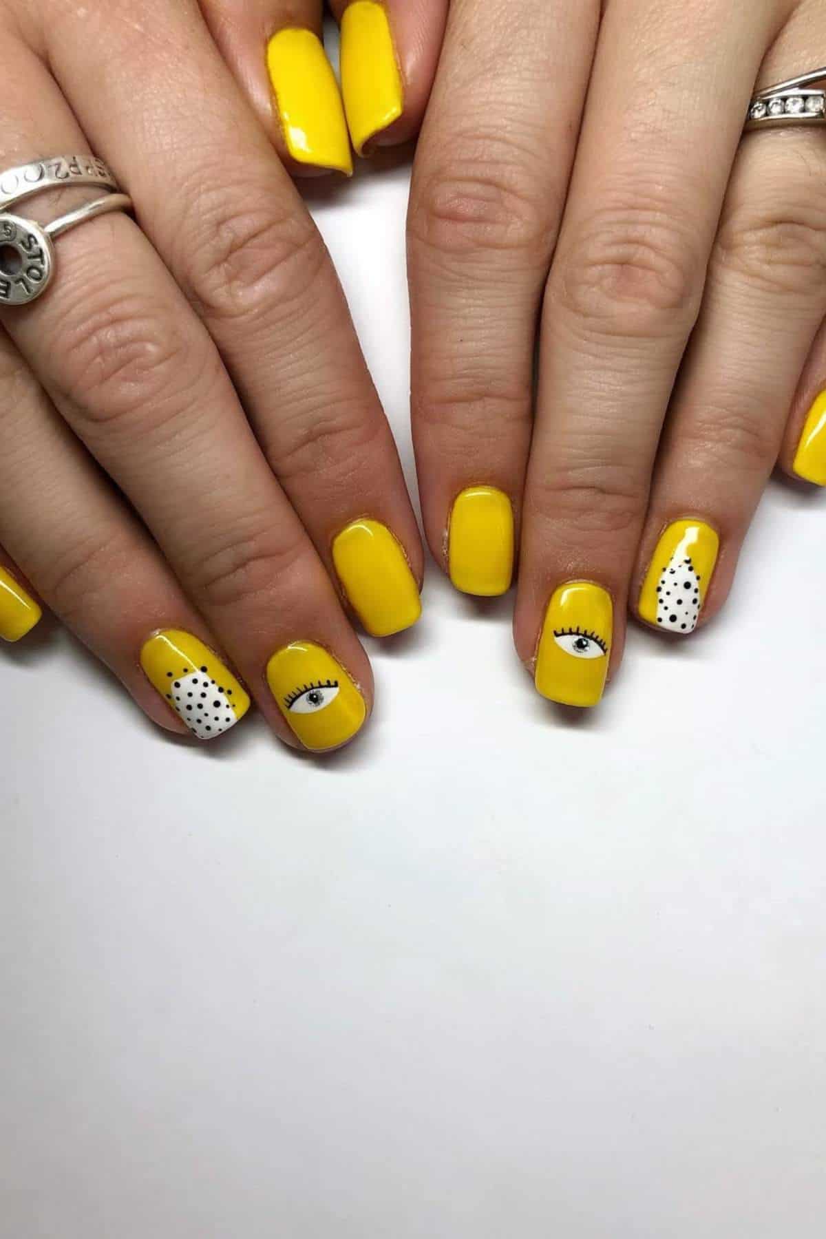 Hands with yellow nail polish; some nails feature black and white polka dots, and two nails have an eye design.