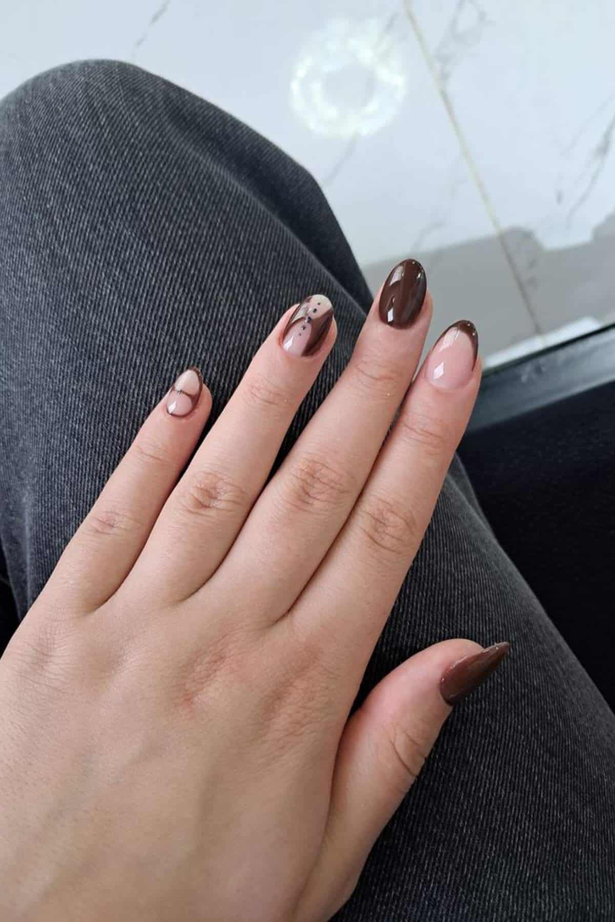 A hand with manicured nails in various shades of brown and nude, featuring abstract patterns and French tips, resting on a leg in dark denim pants.