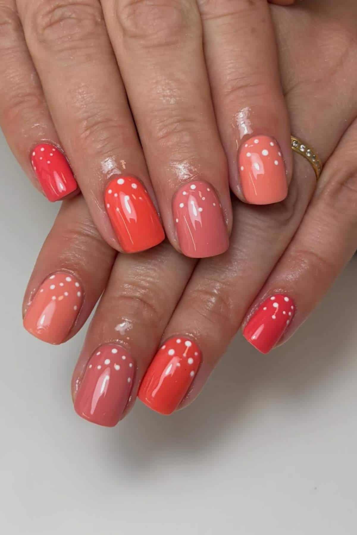 Hands with short, rounded nails painted in various shades of pink and coral, each nail decorated with small white polka dots near the cuticles.