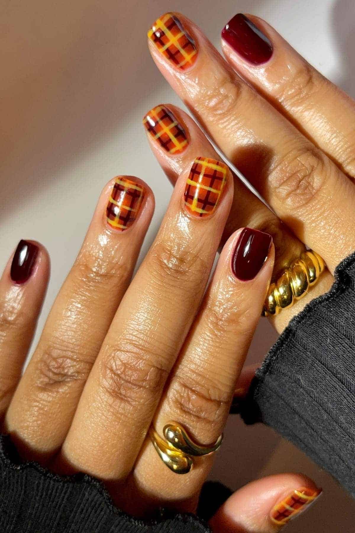 Hands with burgundy and orange plaid patterned nail art, wearing gold rings and a black long sleeve top.