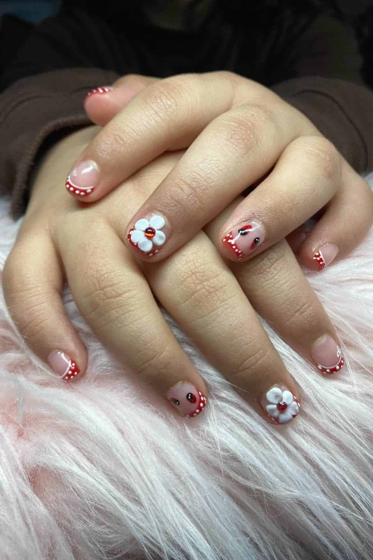 Hands with manicured nails featuring red polka dot French tips, white 3D flowers, and small cherry designs, resting on a fluffy pink surface.