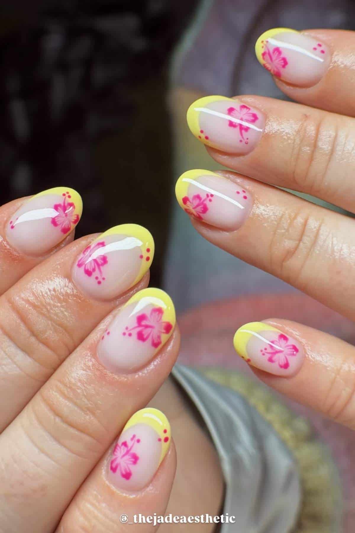 Hands with short, almond-shaped nails featuring yellow French tips and pink floral designs with small red dots on a nude base.