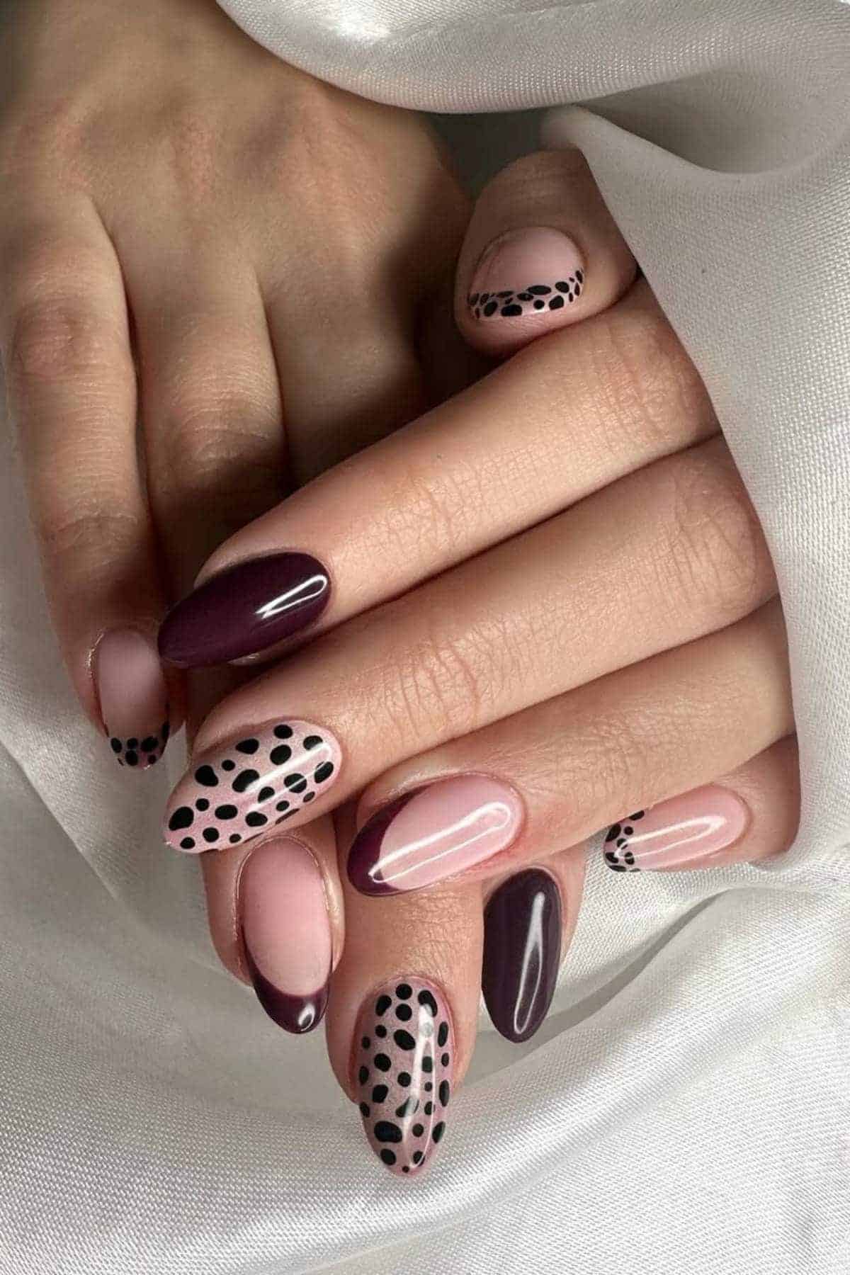 A hand with almond-shaped nails painted in shades of mauve, burgundy, and pale pink, featuring polka dot and French tip designs.