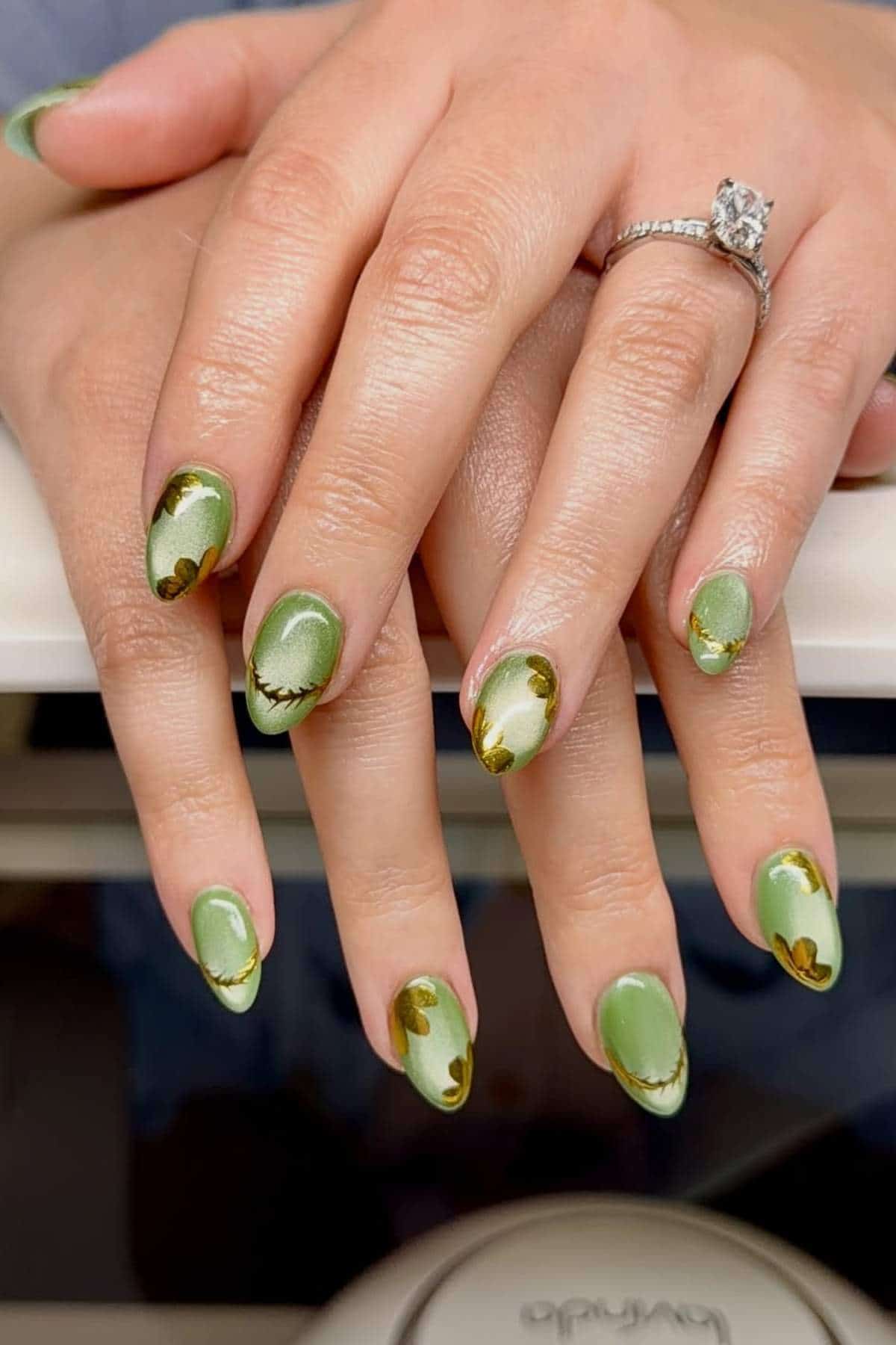 Hands with almond-shaped nails painted in green with gold and dark green abstract accents; one hand wears a silver ring with a large clear stone.