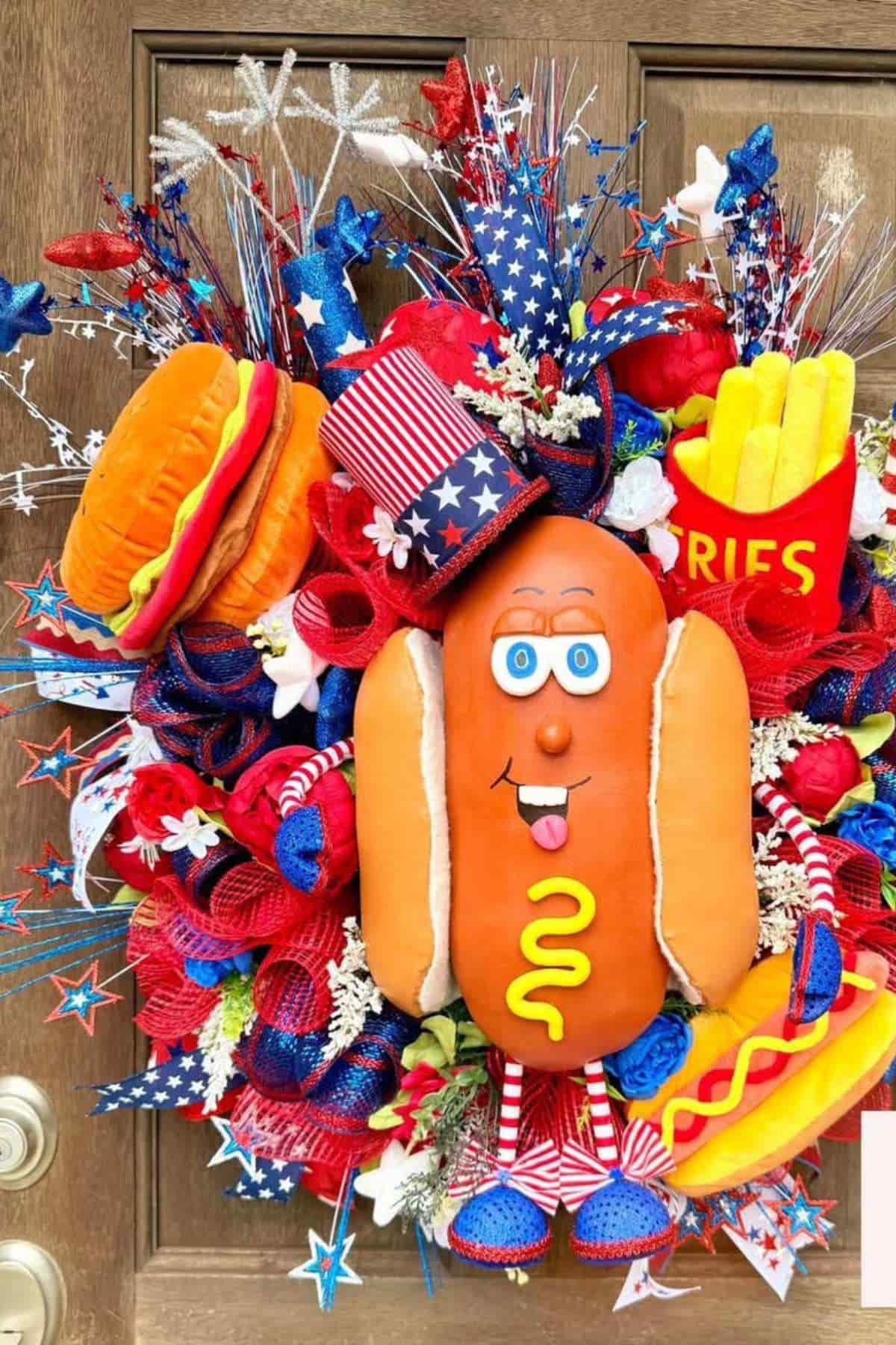 A festive wreath decorated with plush hot dogs, fries, a hamburger, red, white, and blue ribbons, and stars, displayed on a wooden door for a patriotic celebration.