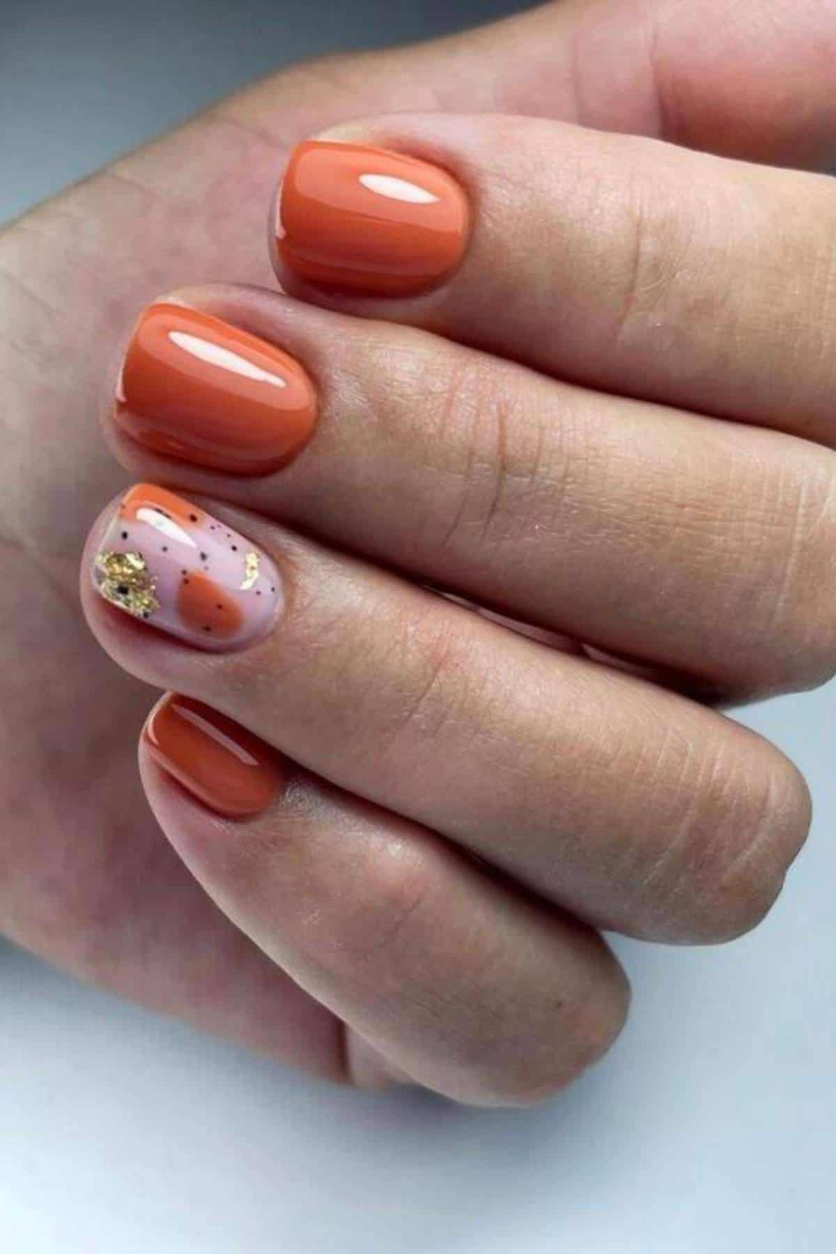 A hand with short, square-shaped nails painted orange, except the ring finger, which has abstract nail art in orange and gold accents on a nude base.