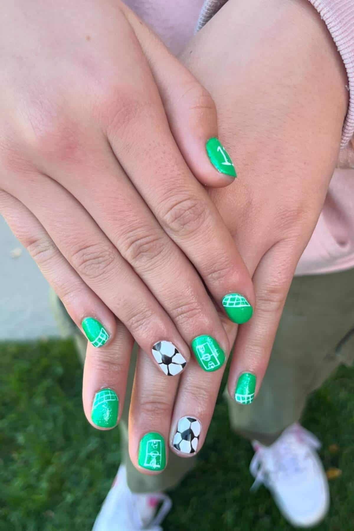Hands with green-painted nails featuring white soccer goal designs and a black-and-white soccer ball pattern on the ring fingernails.