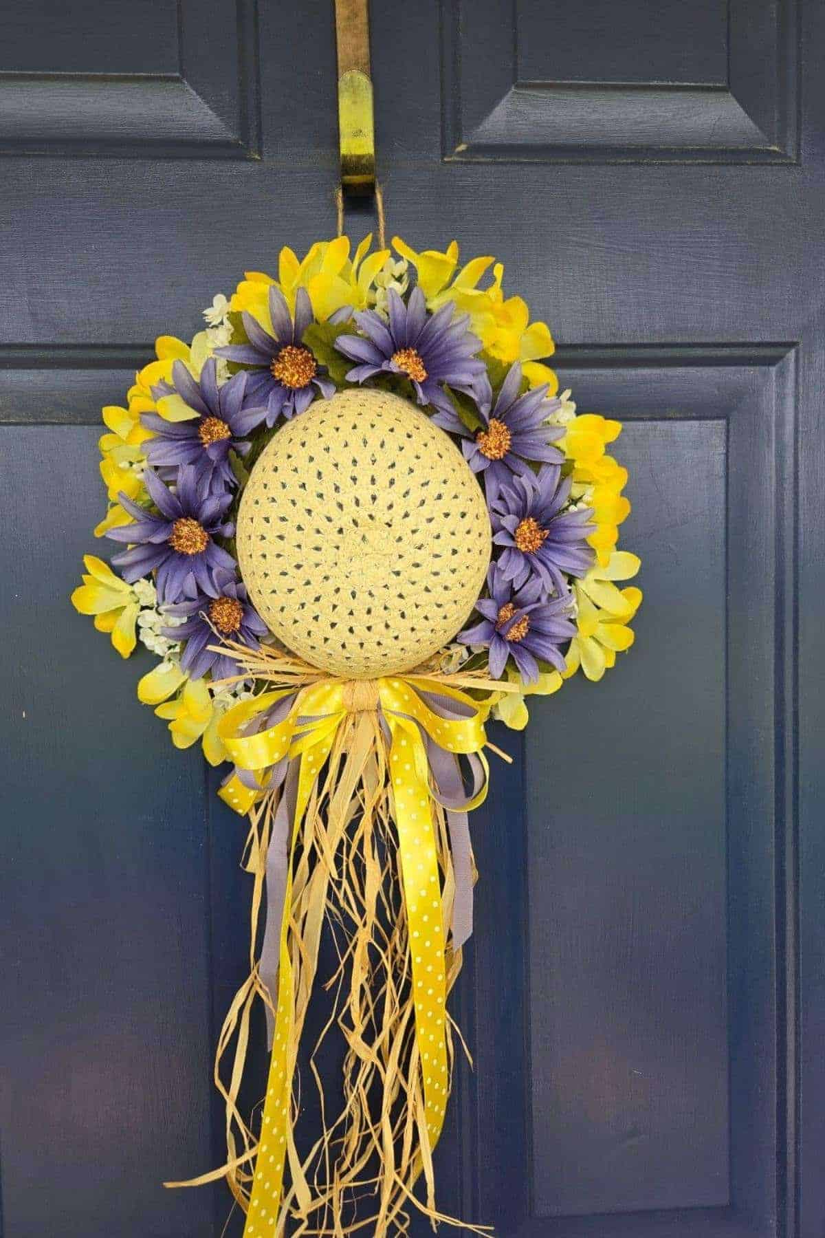 A round door wreath with yellow and purple flowers, a straw hat in the center, and yellow and purple ribbons hanging down, displayed on a dark blue door.