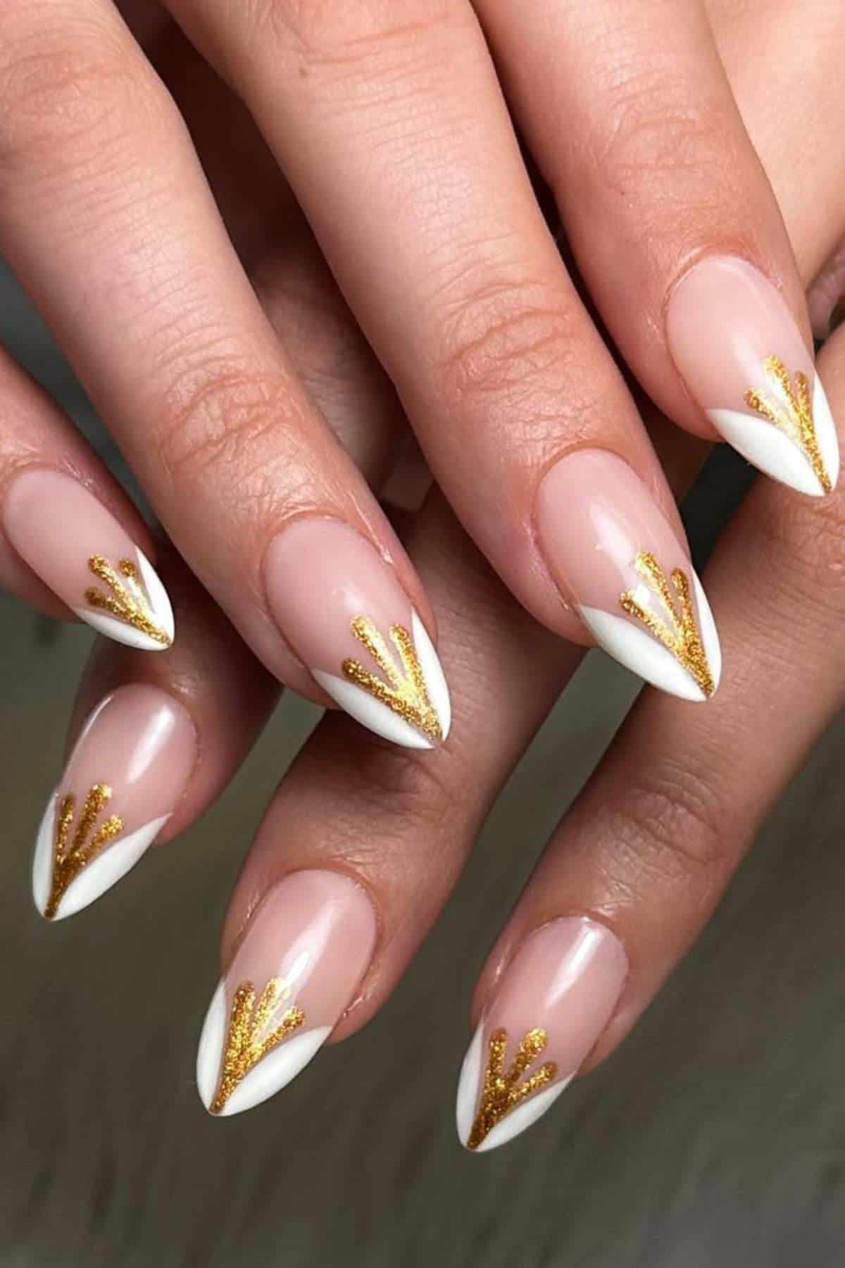 Close-up of hands with almond-shaped nails featuring nude polish, white tips, and gold glitter accents in a geometric design.