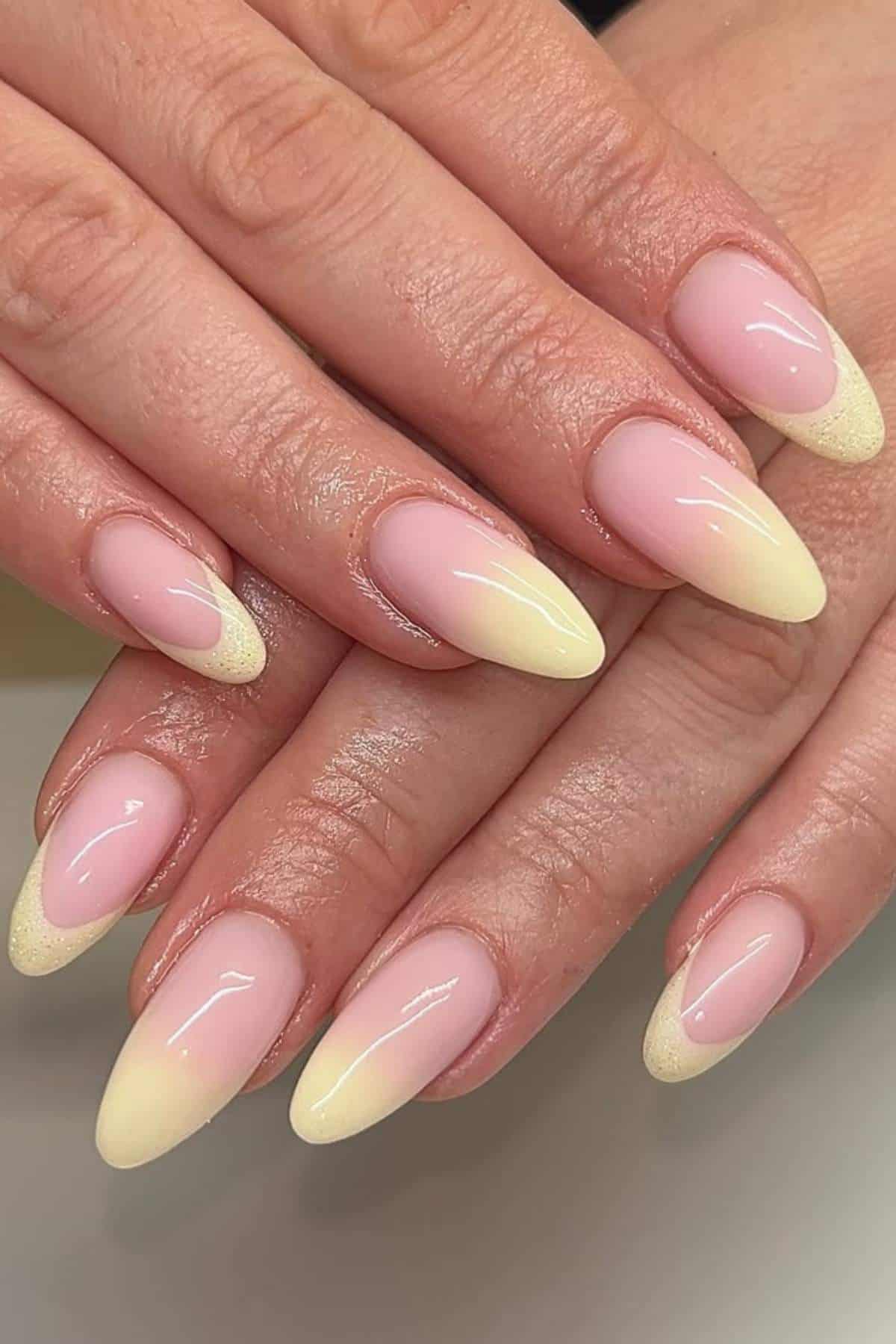 Hands with almond-shaped nails featuring a light pink base and gradient yellow tips, displayed against a neutral background.