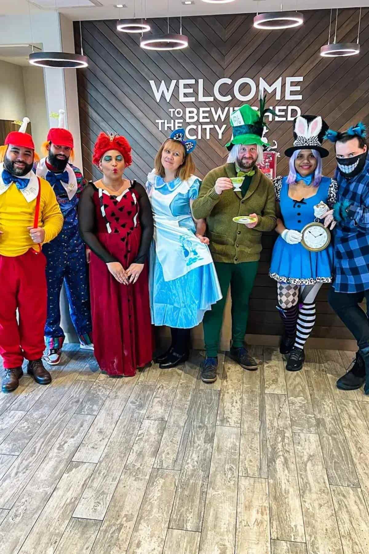 A group of six adults in colorful Alice in Wonderland costumes pose in front of a "Welcome to Bewley" sign on a wood-paneled wall.