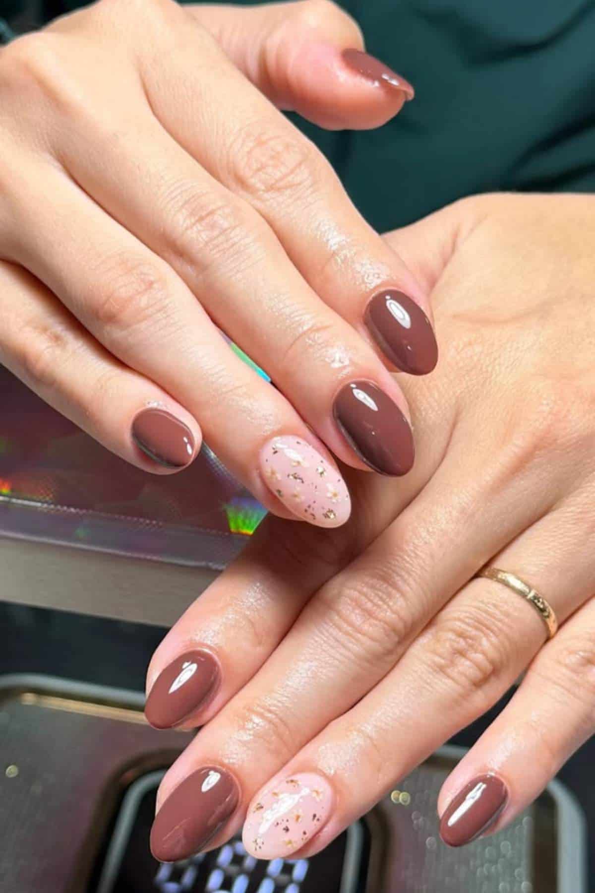 A hand with almond-shaped nails painted in shades of brown and pink, featuring one accent nail with a nude base and gold flakes.