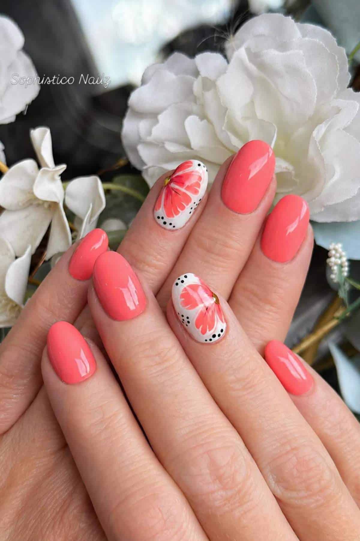 A hand with glossy coral pink nails, featuring an accent nail with a red butterfly and black dot design, is held against white artificial flowers.