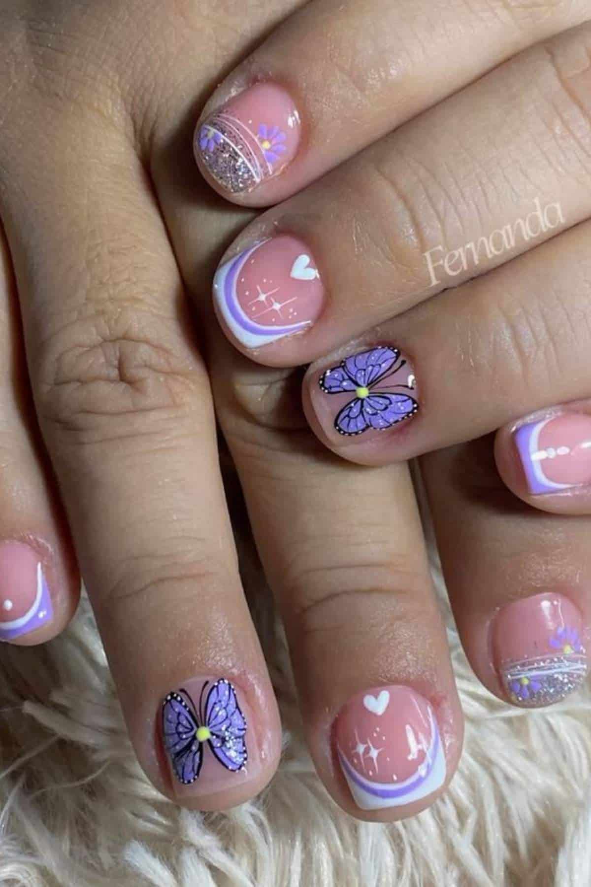 Close-up of hands with short nails featuring pastel purple and white nail art, including butterflies, hearts, stars, and glitter accents.