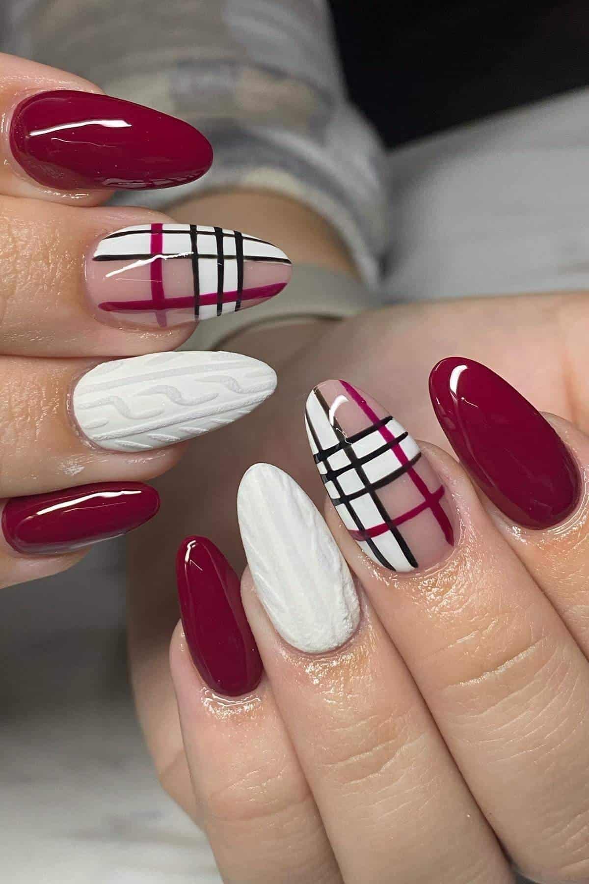 A hand with almond-shaped nails featuring burgundy, white, and black plaid designs, solid burgundy polish, and white textured knit patterns.