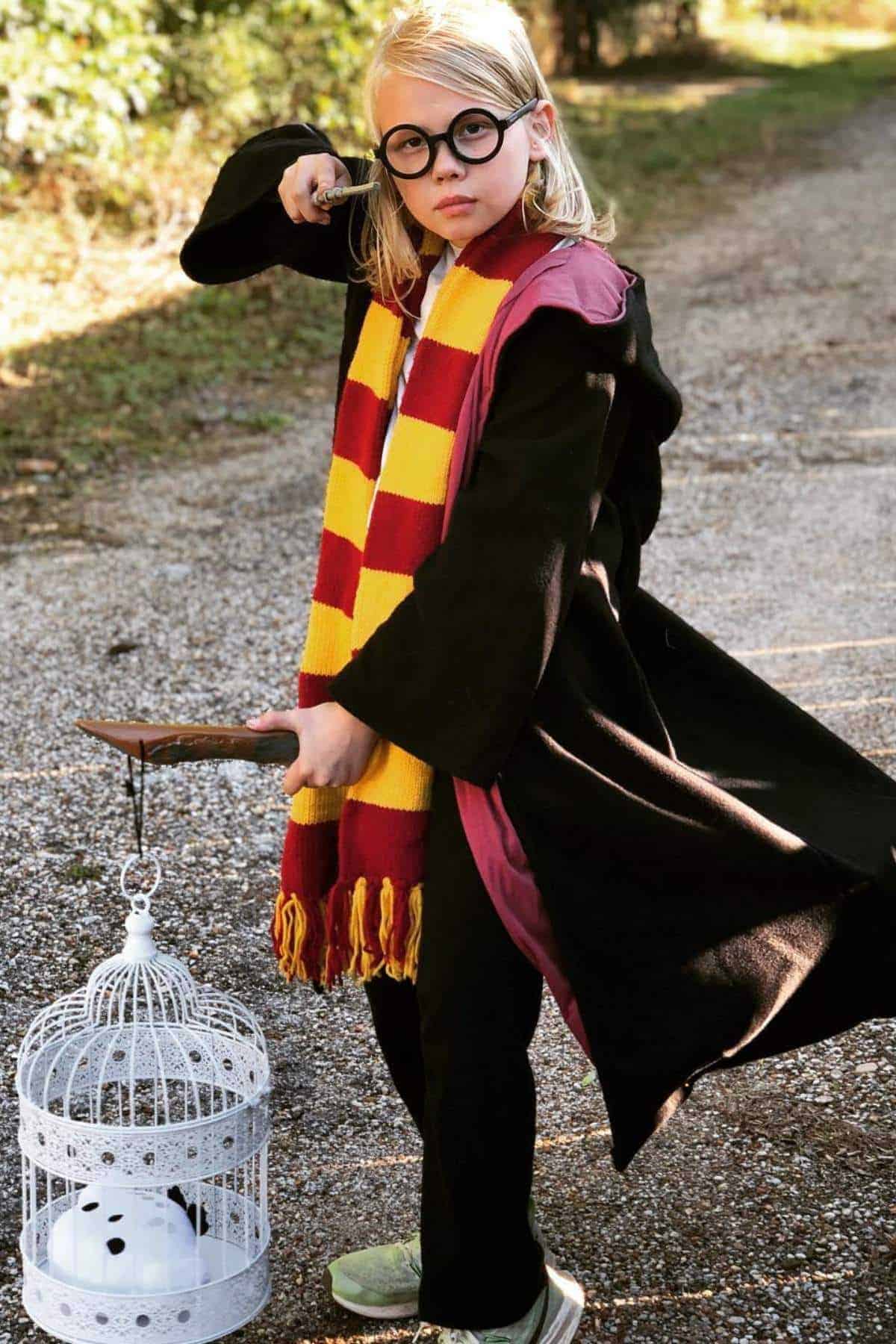 A child dressed in a black robe, red and yellow scarf, and round glasses holds a wand and a white birdcage while standing on a gravel path outdoors.