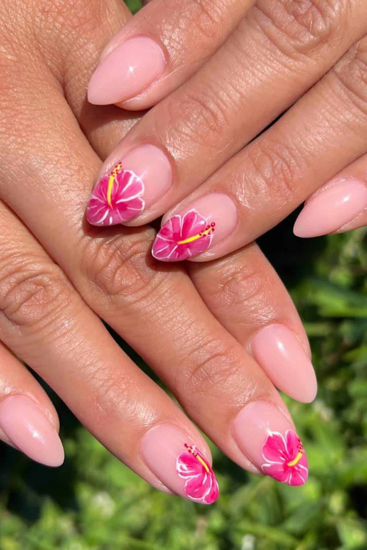 Hands with almond-shaped, nude-colored nails. Two nails on each hand feature detailed pink hibiscus flower nail art. The background is green grass.
