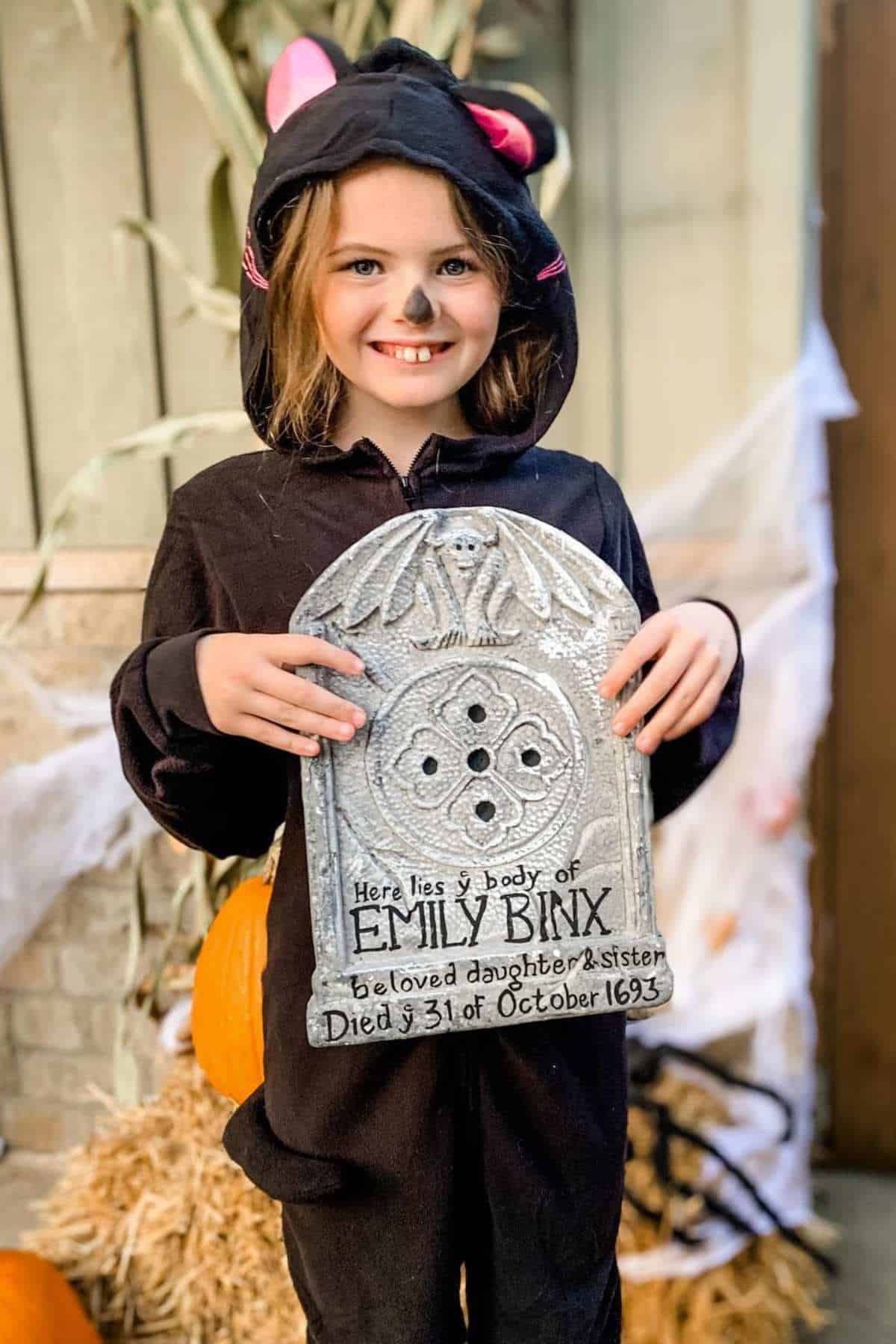 A child in a black cat costume smiles while holding a prop tombstone that reads "Emily Binx" with pumpkins and hay in the background.