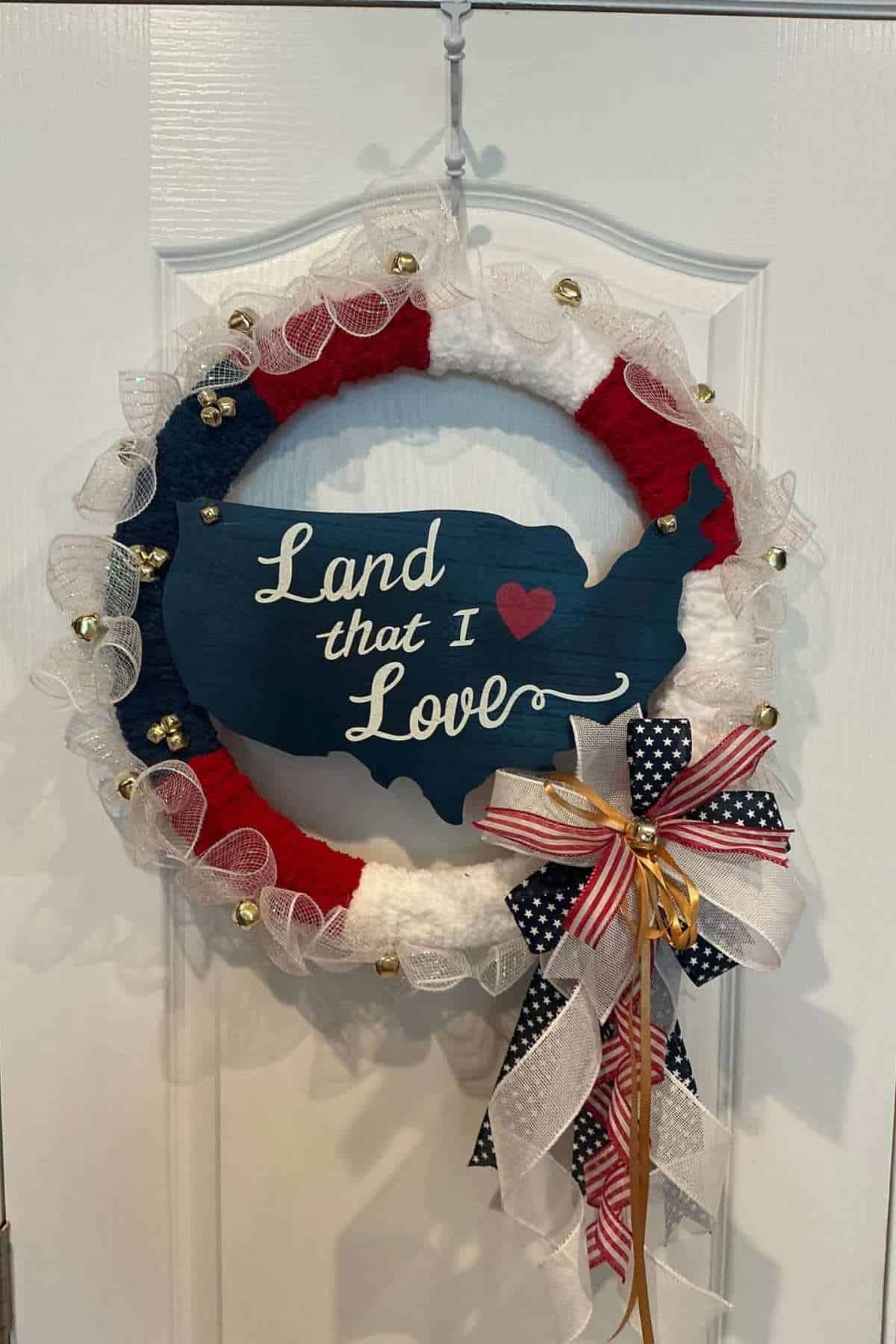 Patriotic wreath with red, white, and blue accents, ribbon bows, and a sign shaped like the U.S. reading “Land that I love” hanging on a white door.