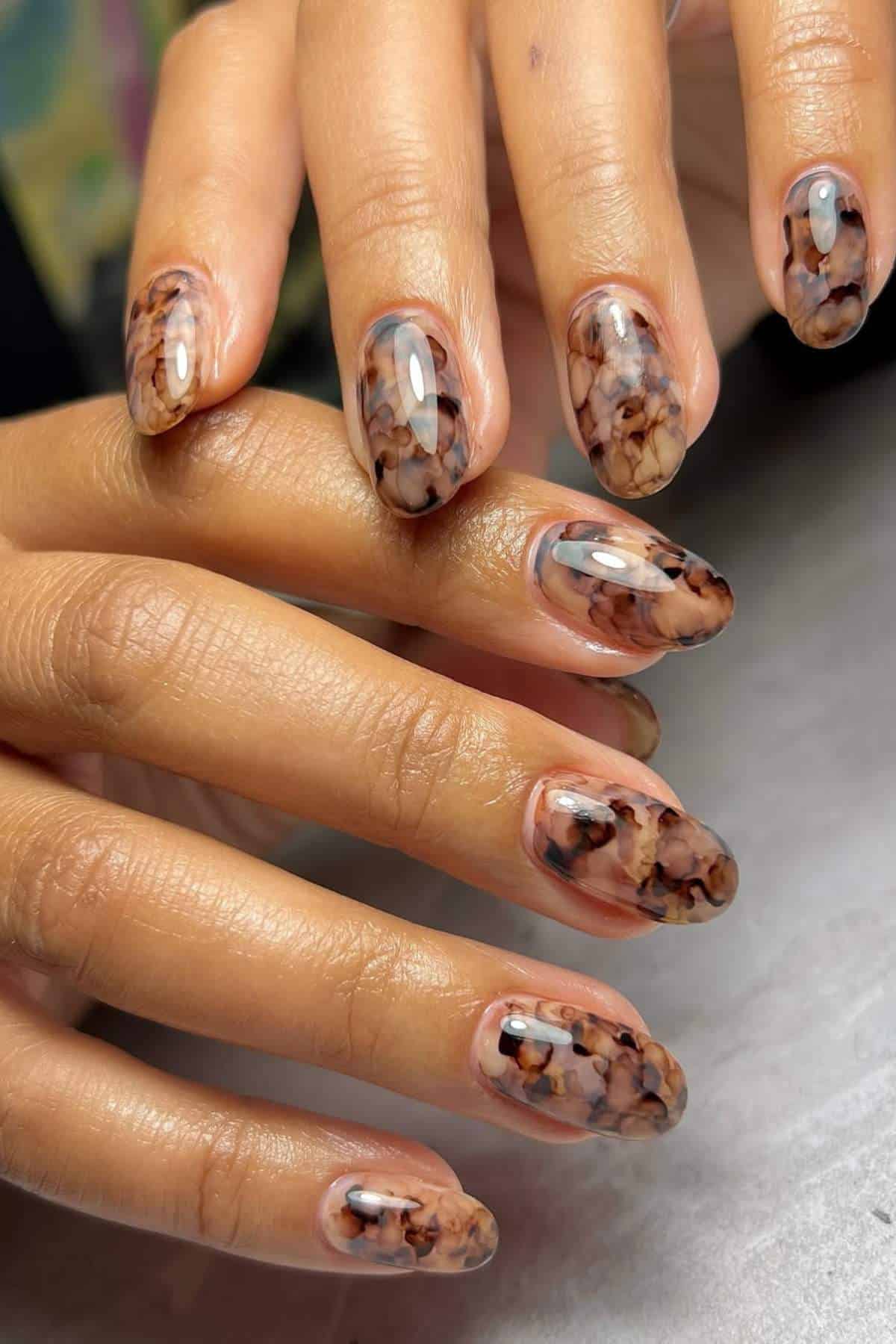 Close-up of hands with almond-shaped nails featuring a marbled brown and black design on a clear polish base.
