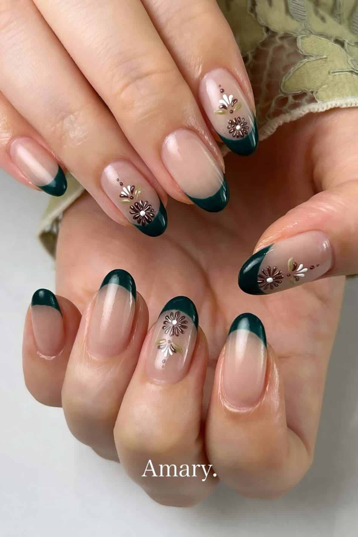 A hand with almond-shaped nails featuring dark green French tips and floral nail art on a nude base.