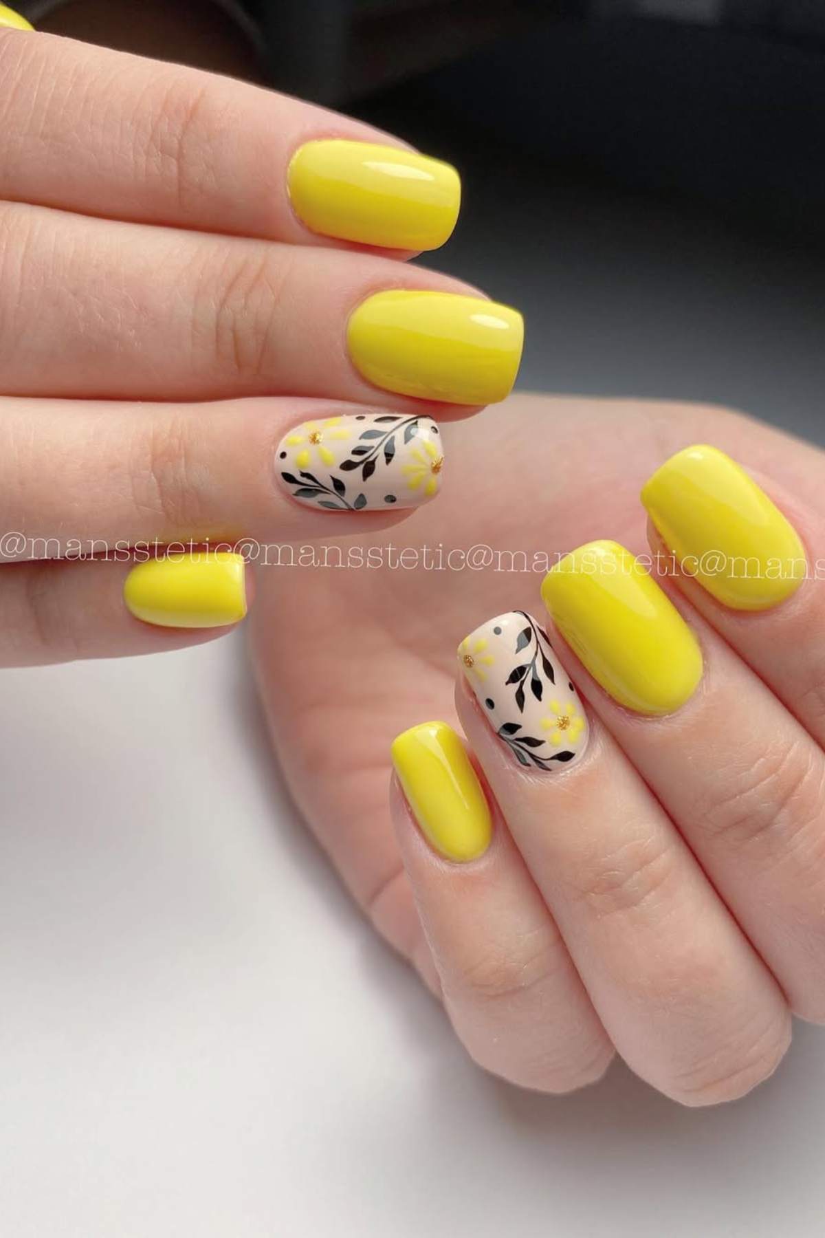 Hands with short, square-shaped nails painted yellow; ring fingers feature floral and leafy black and yellow designs on a nude background.